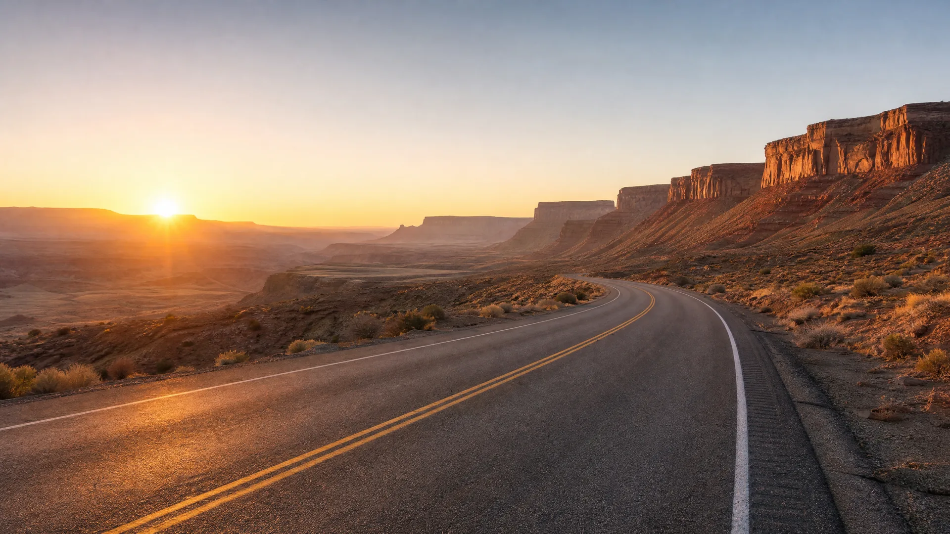 A two-lane asphalt road with double yellow center lines curves through arid desert scrub toward a low sun, with layered sandstone cliffs and mesas on the right.