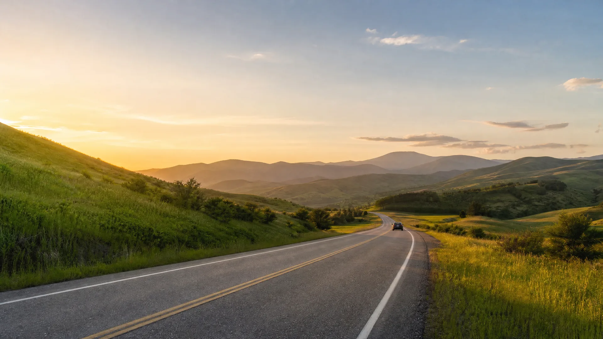 A two-lane paved road with double yellow center lines curves through grassy green hills toward distant mountain ridges at sunrise or sunset, with a single dark car in the distance.
