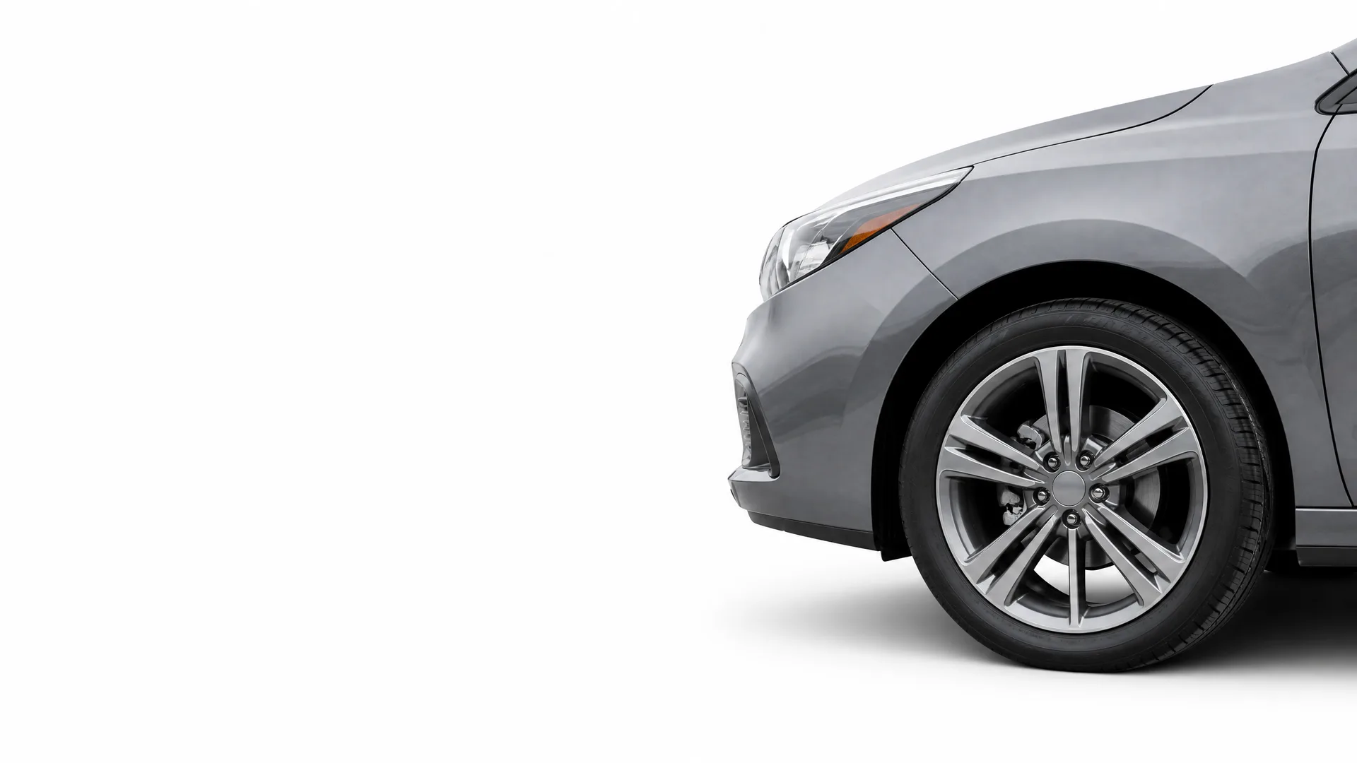 Partial view of the front end of a gray sedan, showing the headlight, bumper, fender and front alloy wheel, against a plain white background.