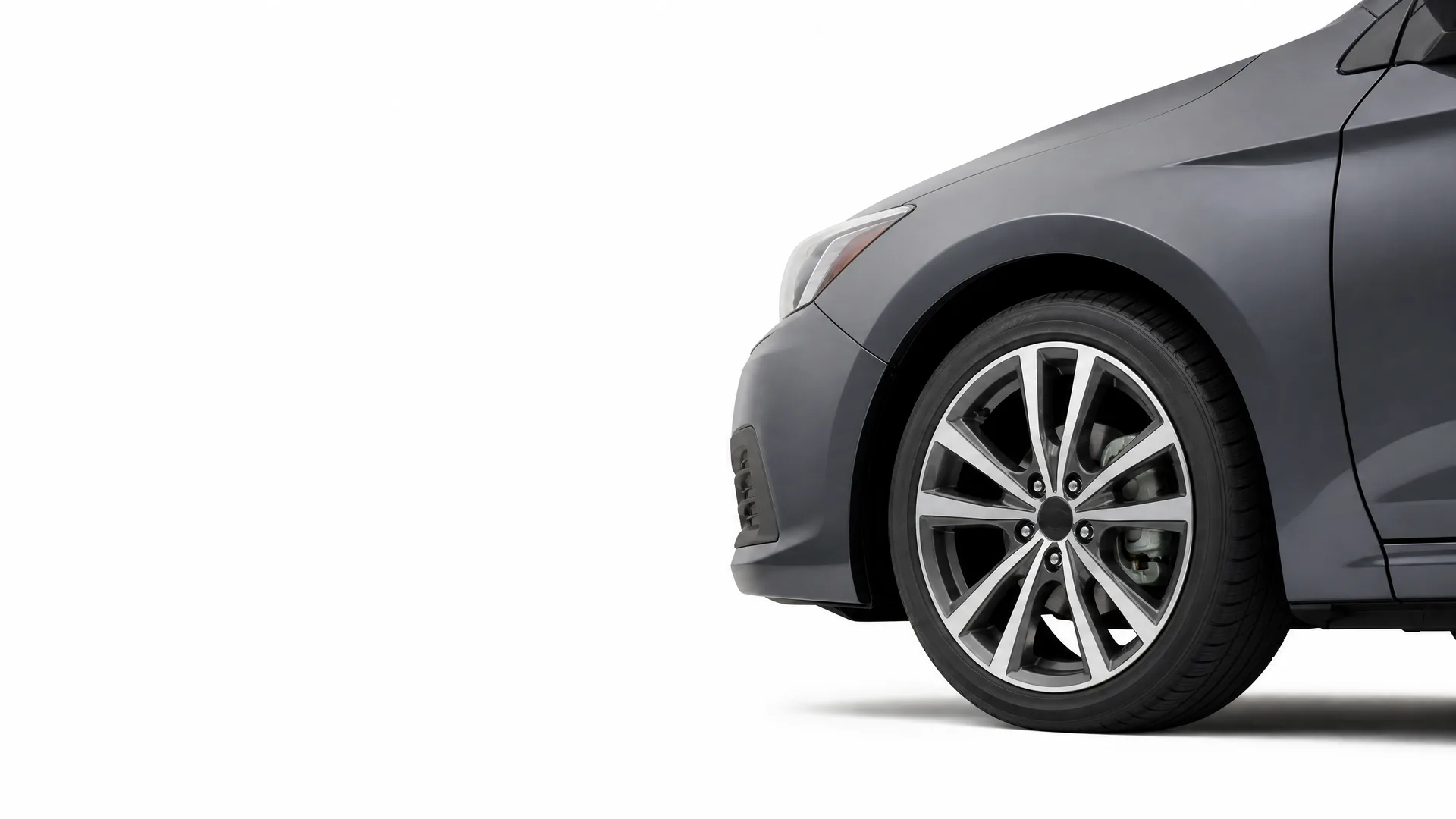 Partial view of the front left side of a dark gray car, showing the front wheel with a multi-spoke alloy wheel, headlight and bumper, against a plain white background.