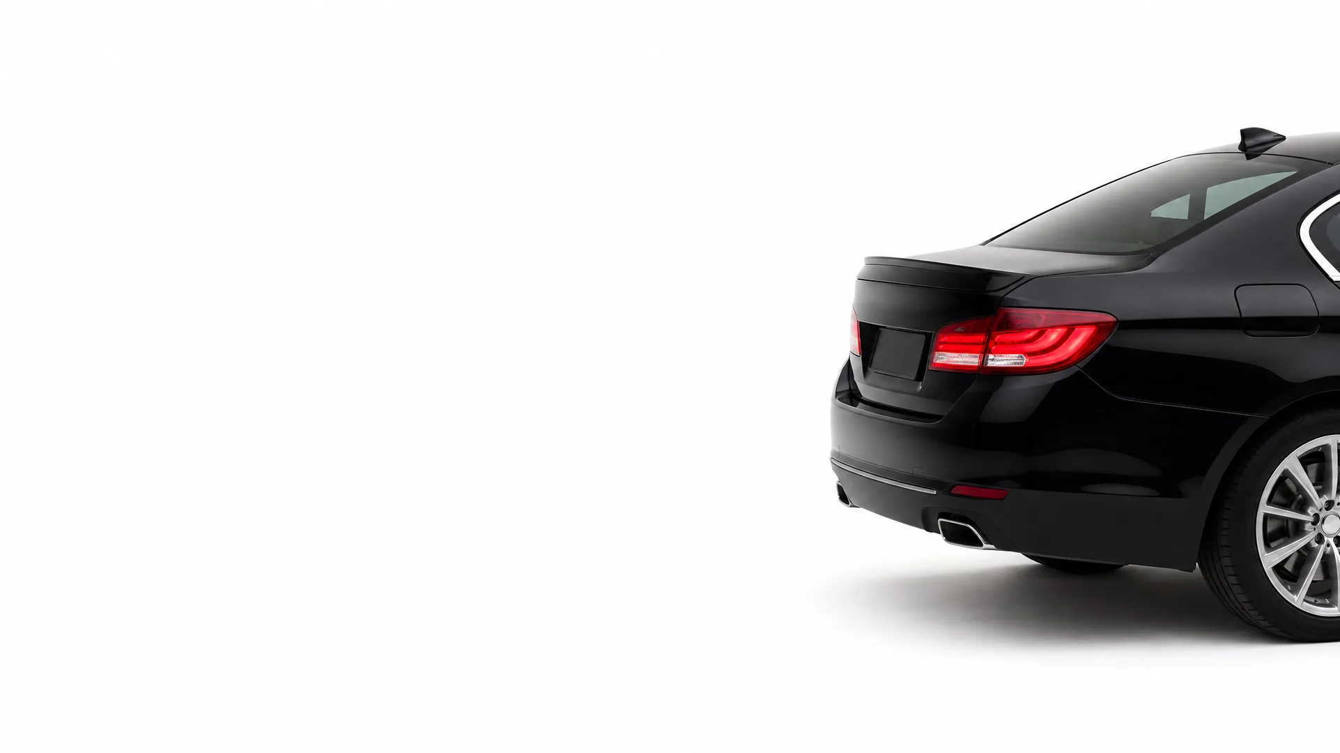Rear three-quarter view of a black sedan against a plain white background, showing the taillight, trunk, exhaust outlet and alloy wheel.