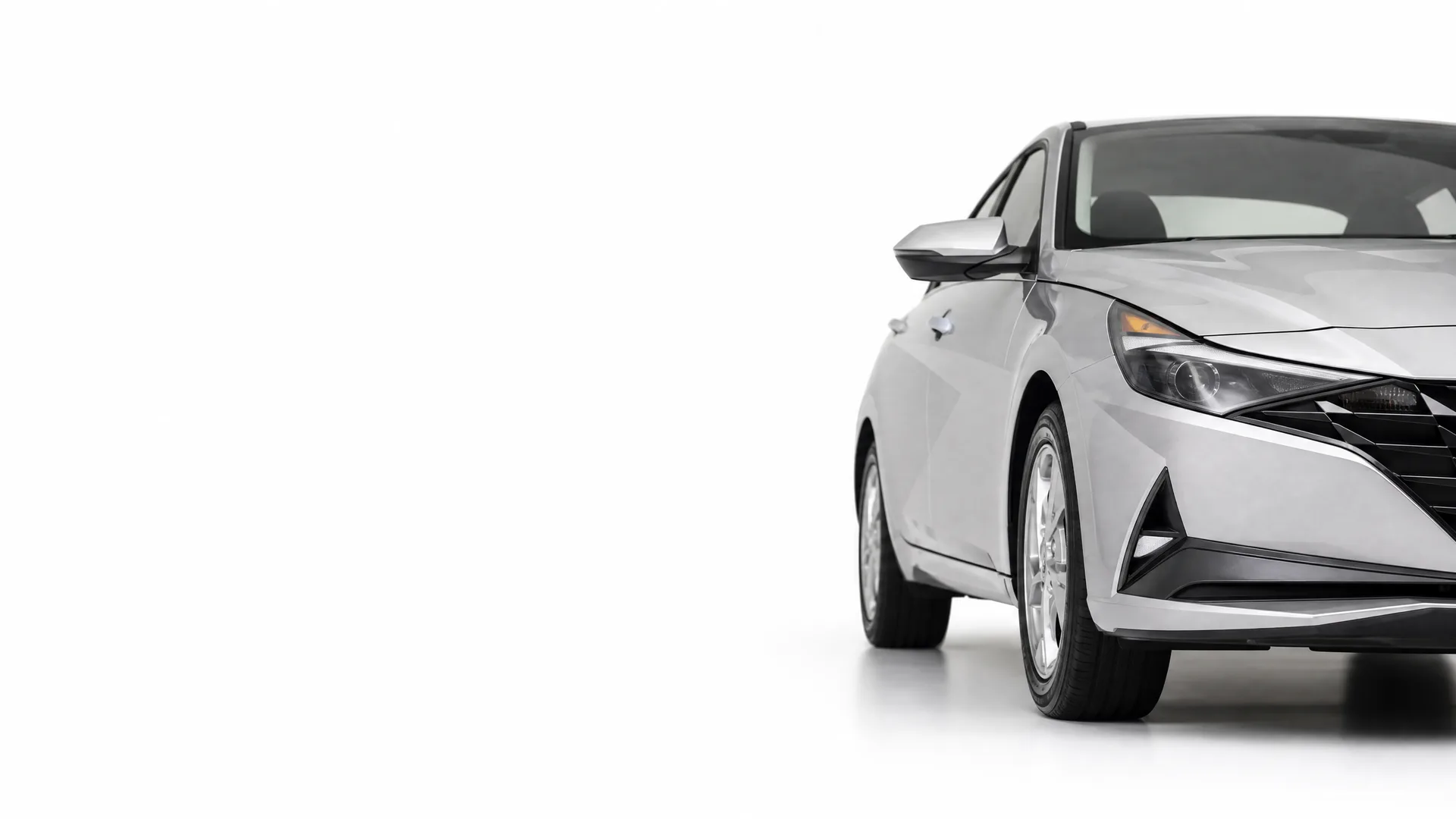 Front half of a silver four-door sedan photographed against a plain white studio background, showing the headlight, front wheel, side mirror and part of the grille.