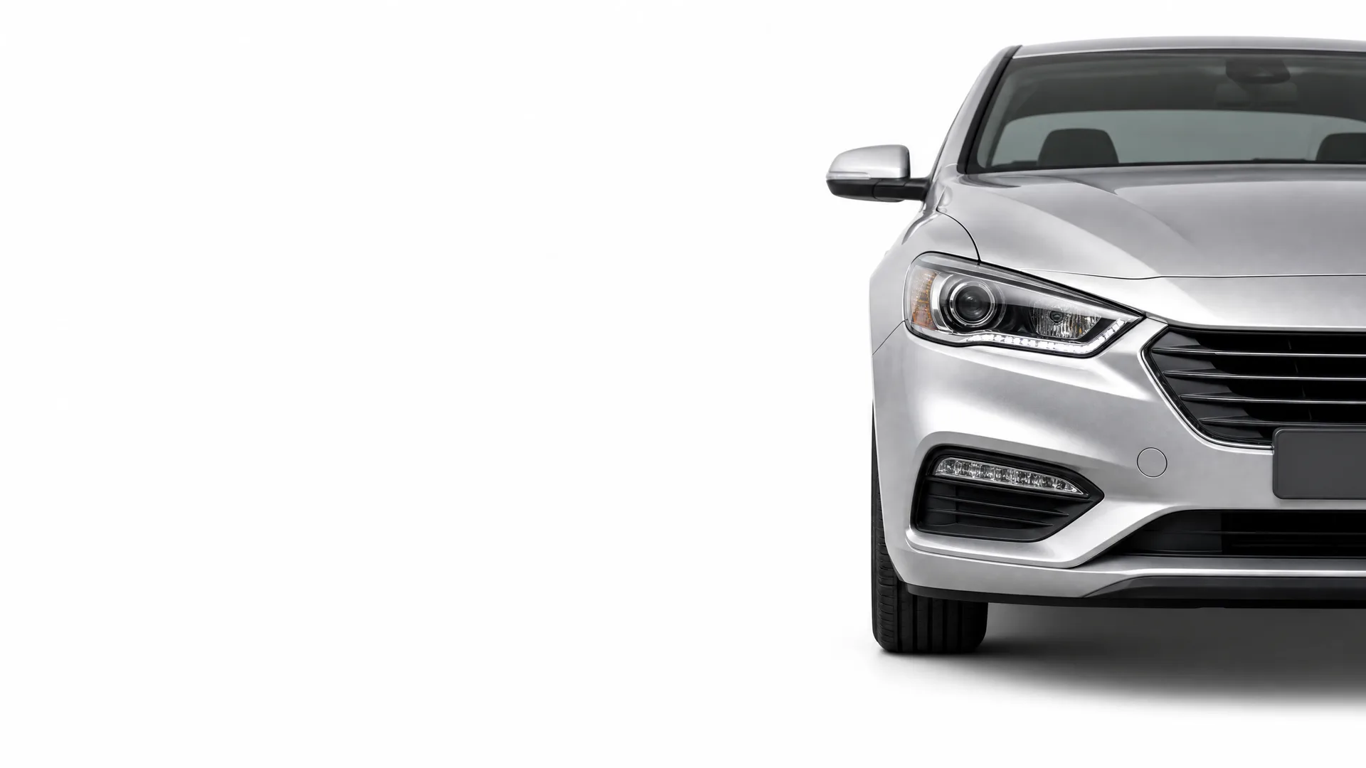 Front left portion of a silver sedan, showing the headlight, grille, bumper and front wheel, against a plain white background.