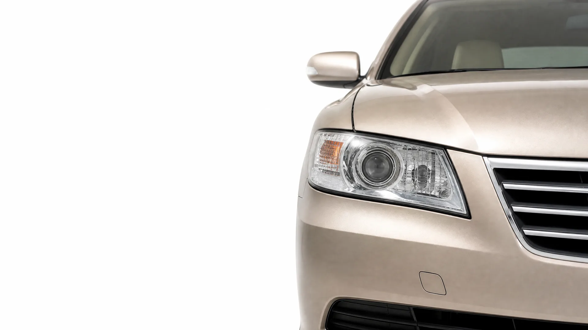 Close-up of the front left corner of a beige sedan, showing the headlight, side mirror, windshield and part of the grille, against a white background.