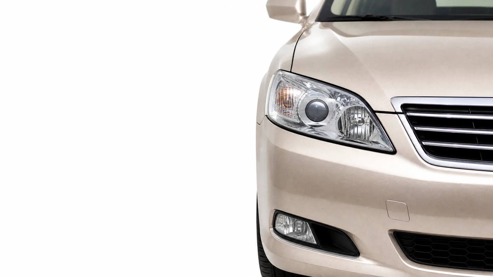 Close-up of the front right corner of a beige sedan, showing the headlight, grille, fog light and bumper against a white background.