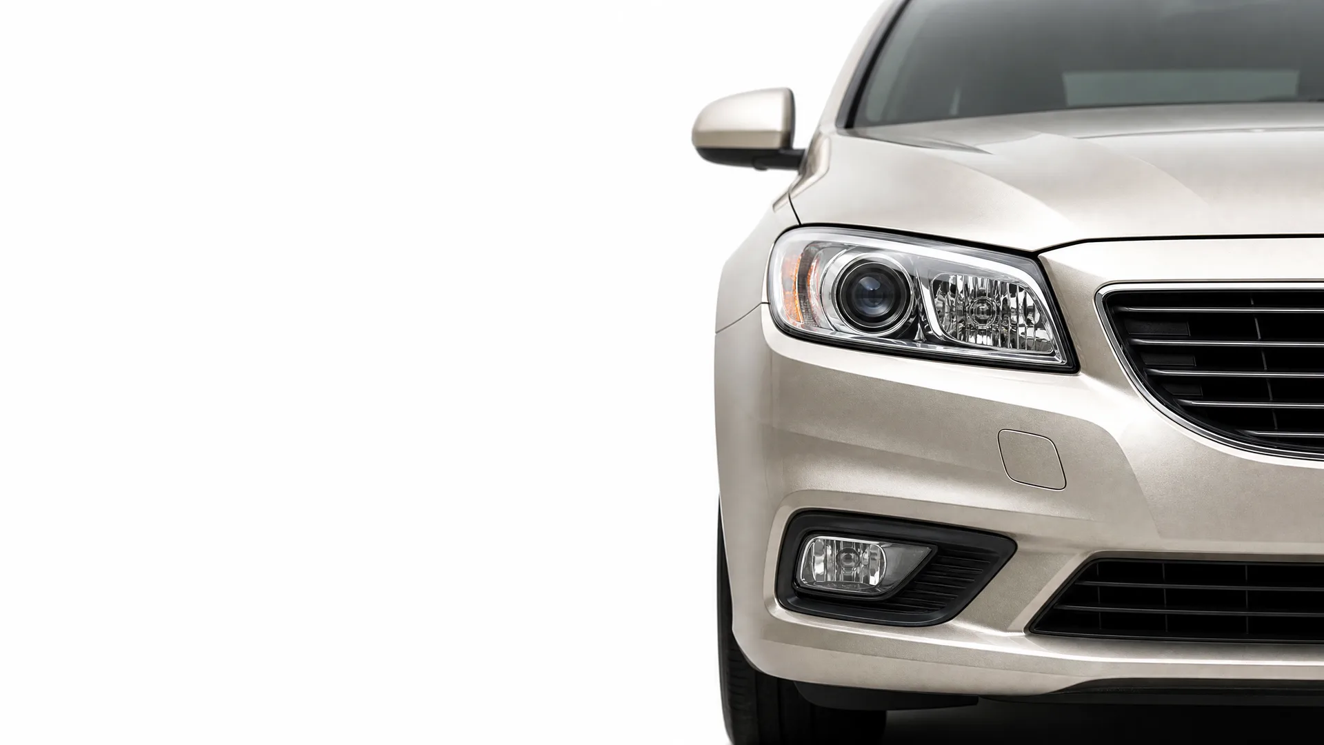 Close-up of the front left corner of a silver-beige car, showing the headlight, fog lamp, grille edge and side mirror, against a plain white background.