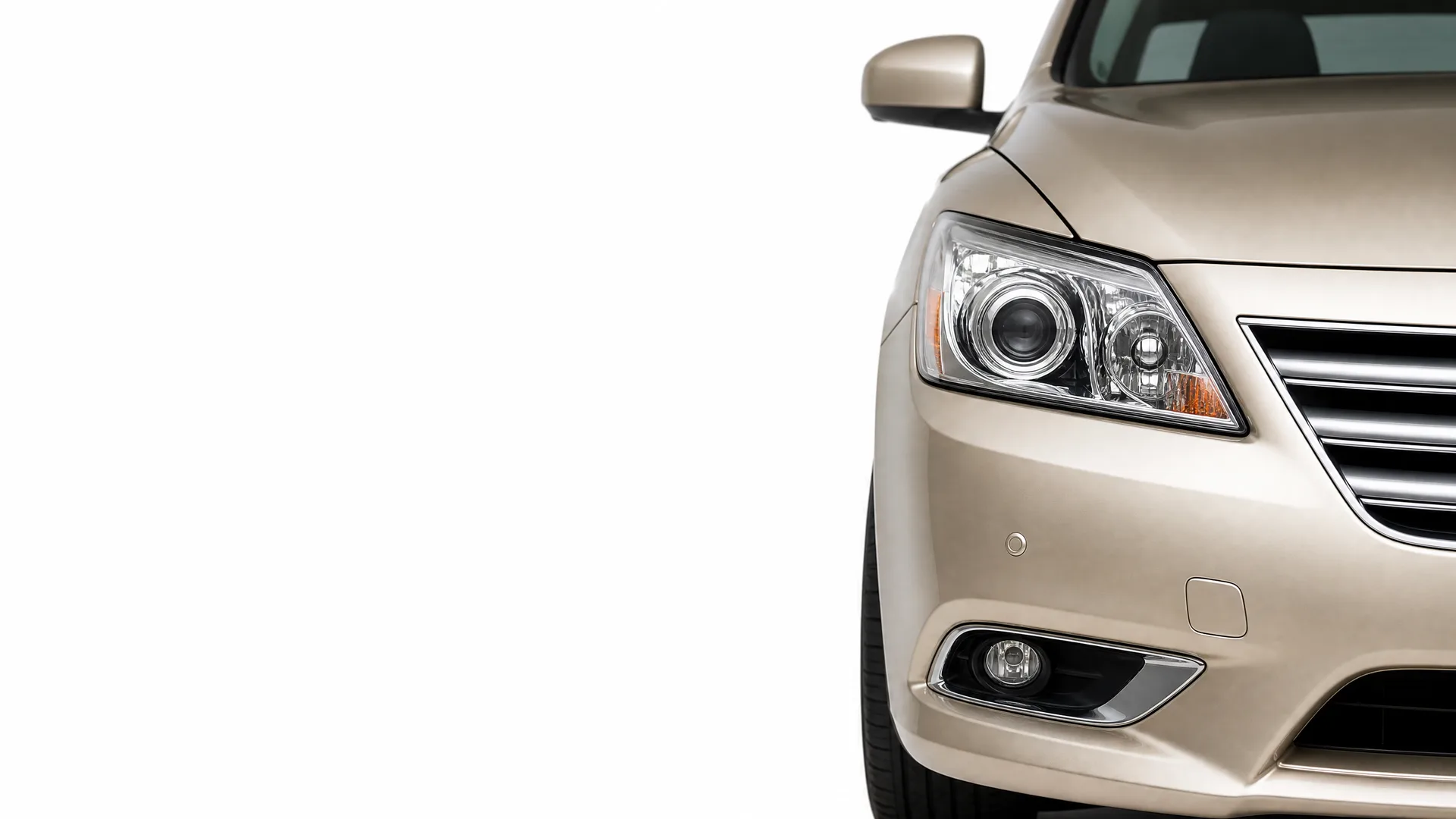 Close-up of the front right corner of a beige sedan, showing the headlight, grille, fog lamp and side mirror against a white background.