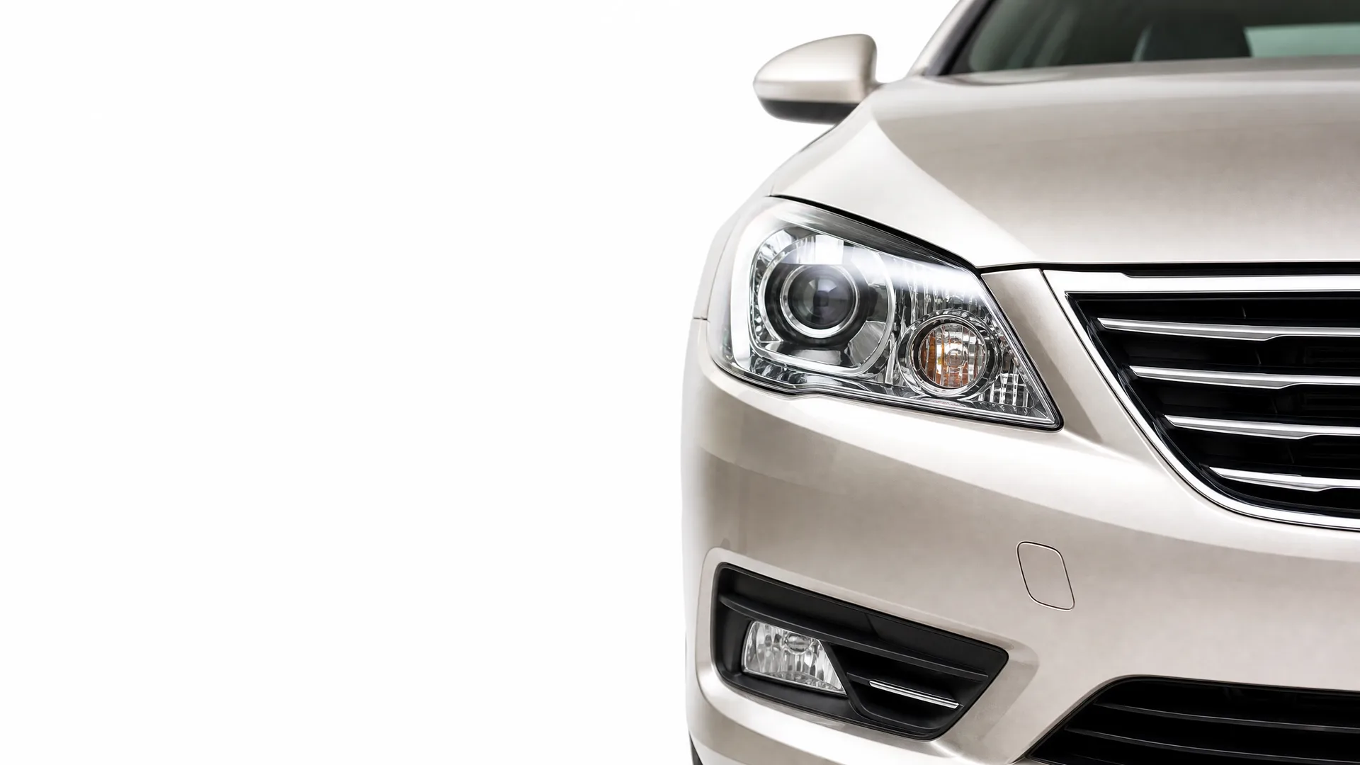 Close-up of the front left corner of a silver-beige car, showing the headlight, grille, fog light and side mirror against a white background.
