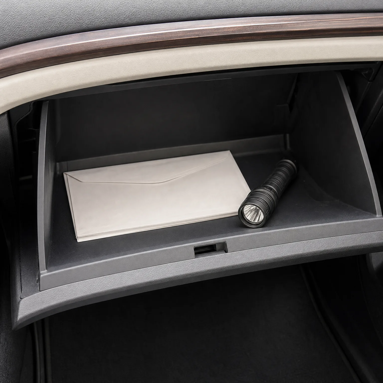 An open car glove compartment containing a white envelope and a small black flashlight, below a dashboard with wood-grain trim.