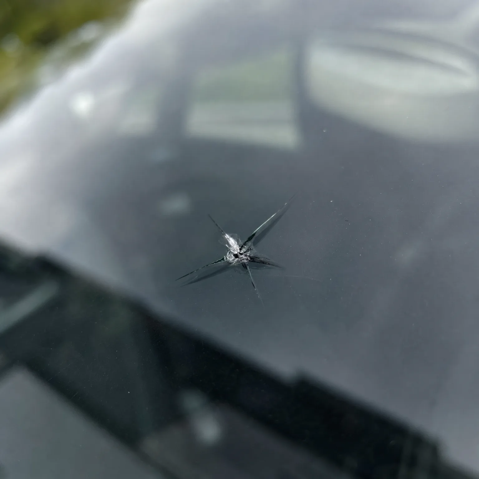 Close-up of a star-shaped chip with radiating cracks in a car windshield, with blurred green foliage and vehicle reflections in the background.
