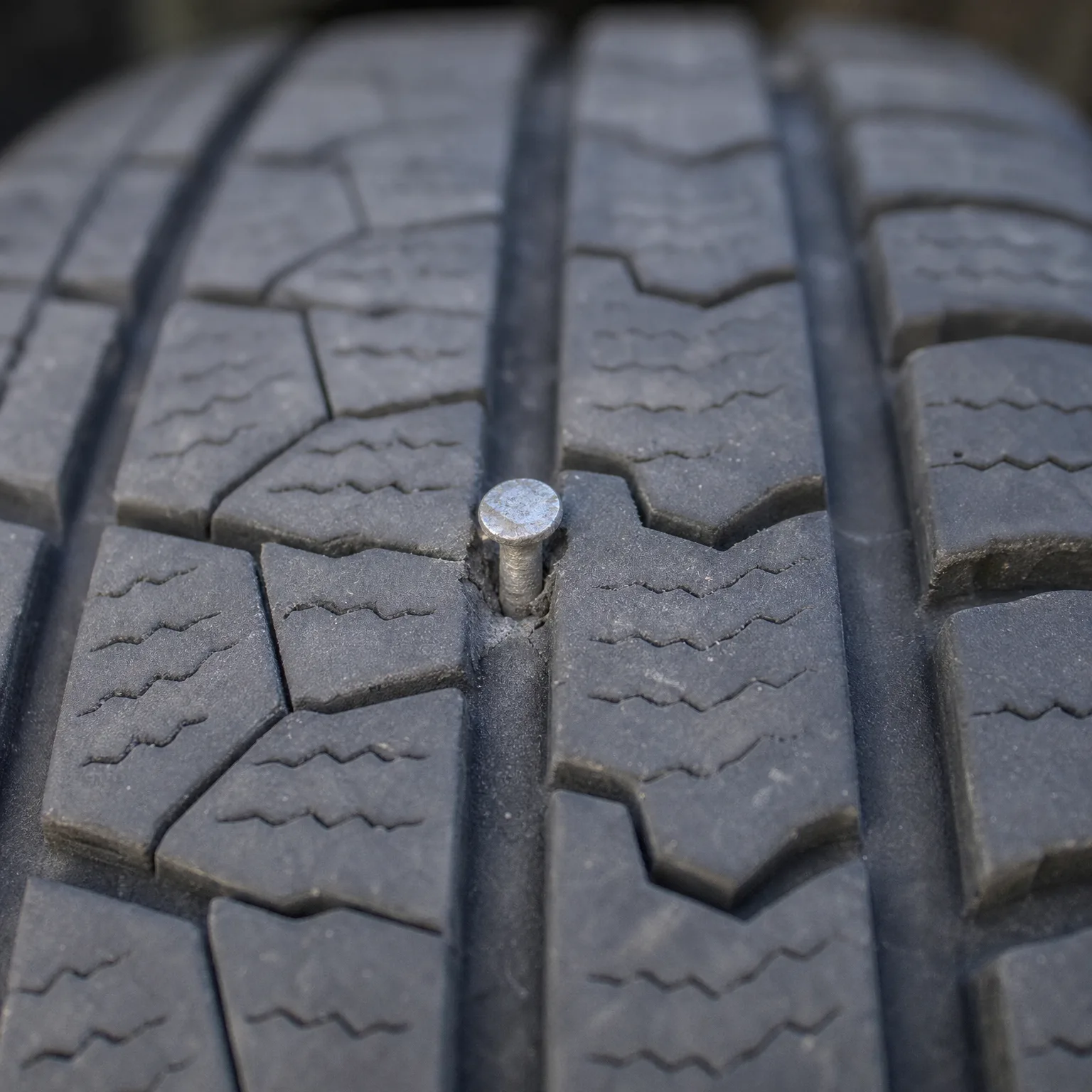 Close-up of a tire tread with a metal nail head embedded beside a longitudinal groove.