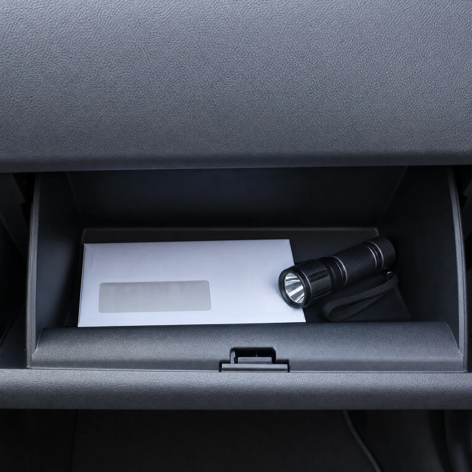 An open car glove compartment containing a white envelope with a clear address window and a black flashlight with a wrist strap.
