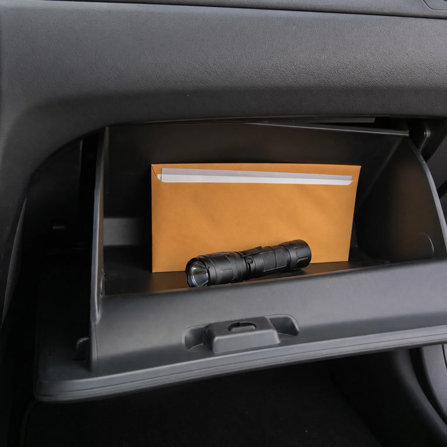 An open car glove compartment containing a black flashlight resting in front of a tan manila envelope.