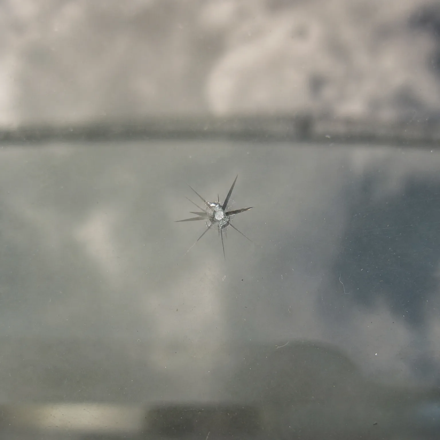 Close-up of a star-shaped chip in glass with radiating cracks, against a blurred light-colored background seen through the pane.
