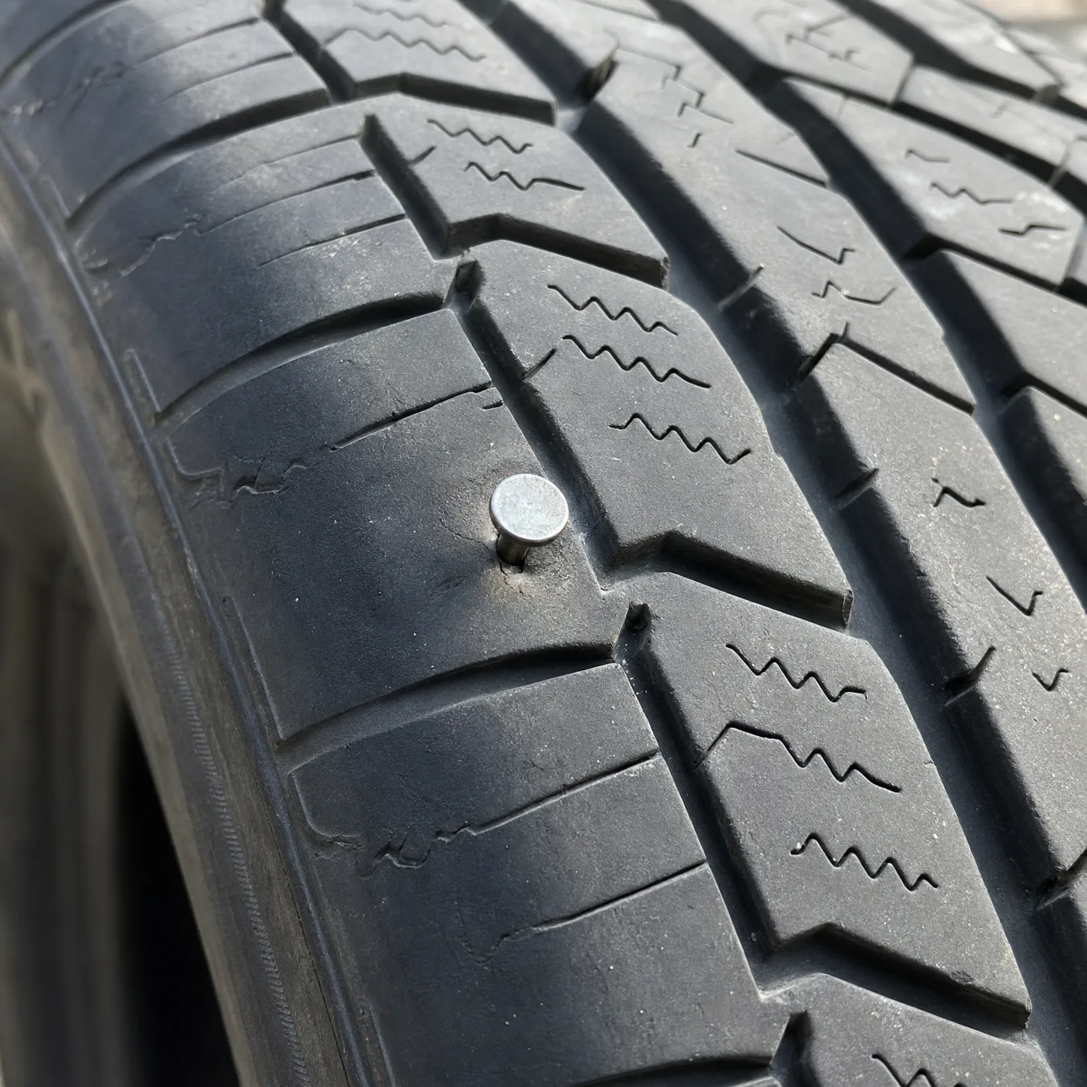 Close-up of a black tire tread with a metal nail head embedded in the rubber between tread blocks.