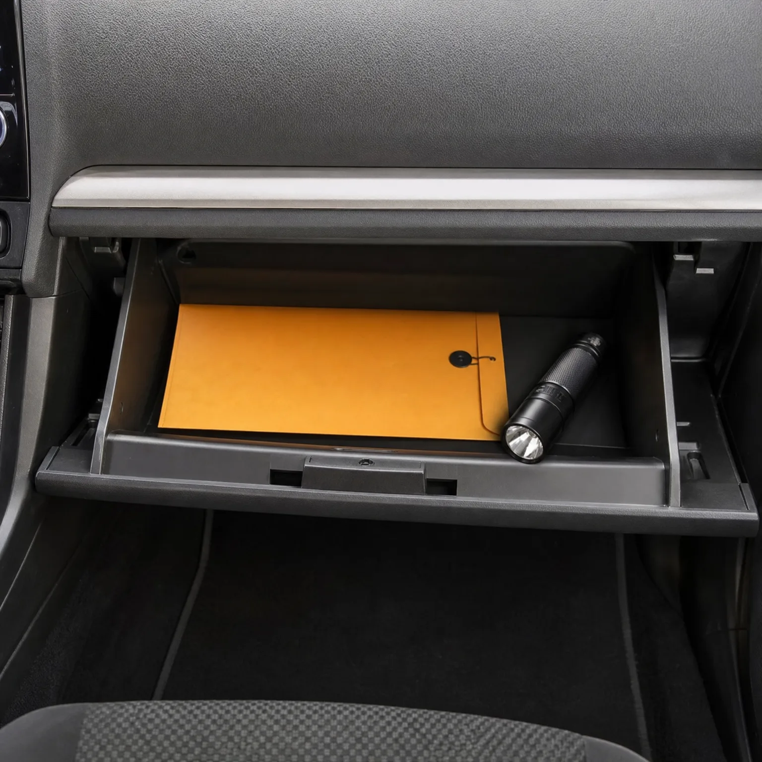 An open car glove compartment containing an orange envelope and a black flashlight, with the dashboard above and floor mat below.