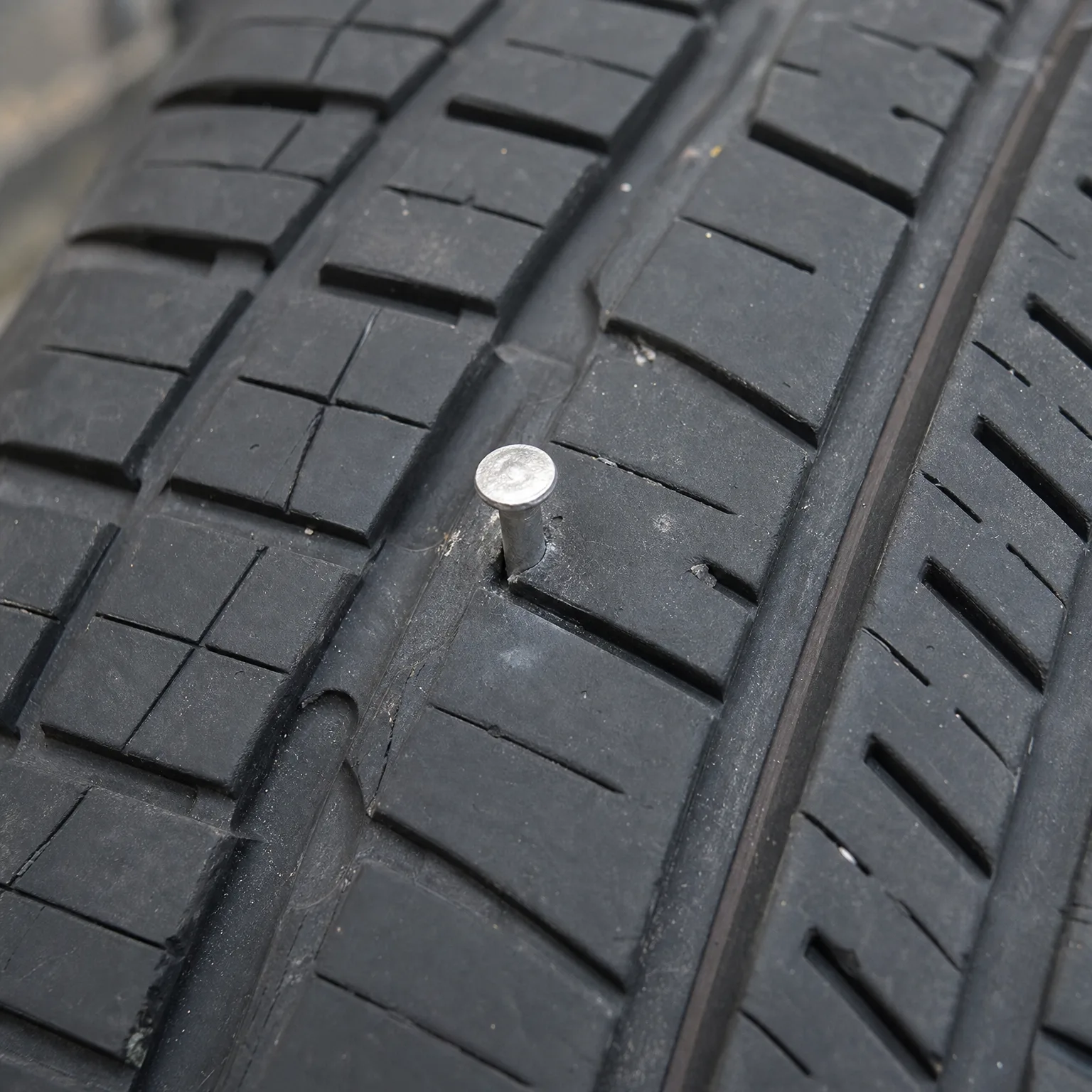 Close-up of a black tire tread with a metal nail head embedded in the rubber between grooves.