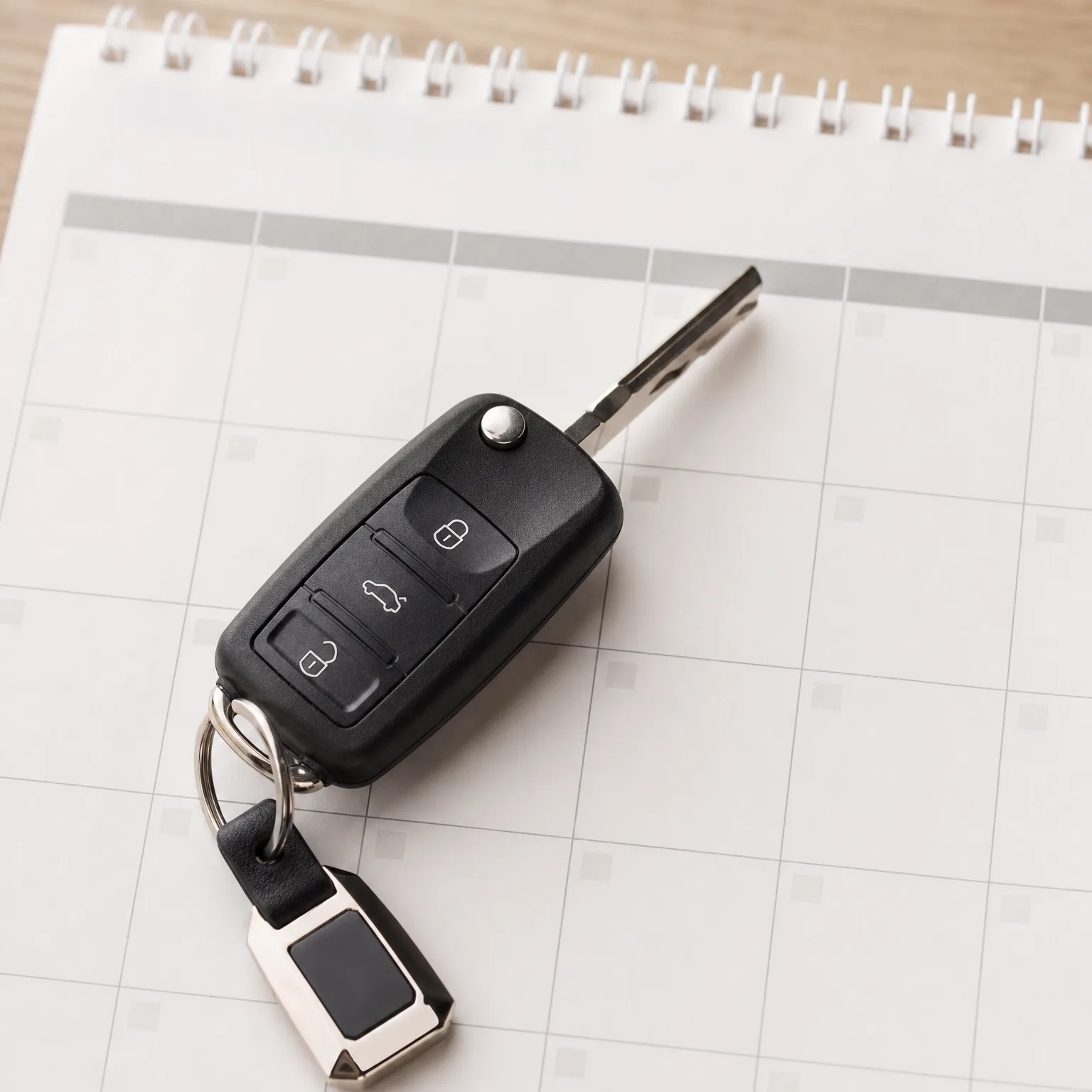 A black folding car key with three buttons and a leather keychain fob rests on an open spiral-bound calendar page on a wooden surface.