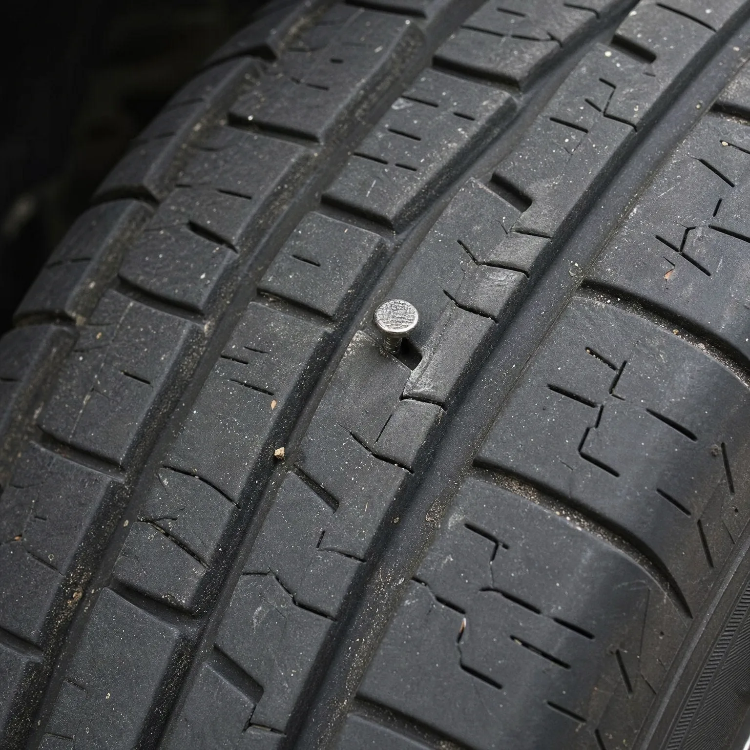 Close-up of a dusty tire tread with a metal screw head embedded in the rubber between grooves.