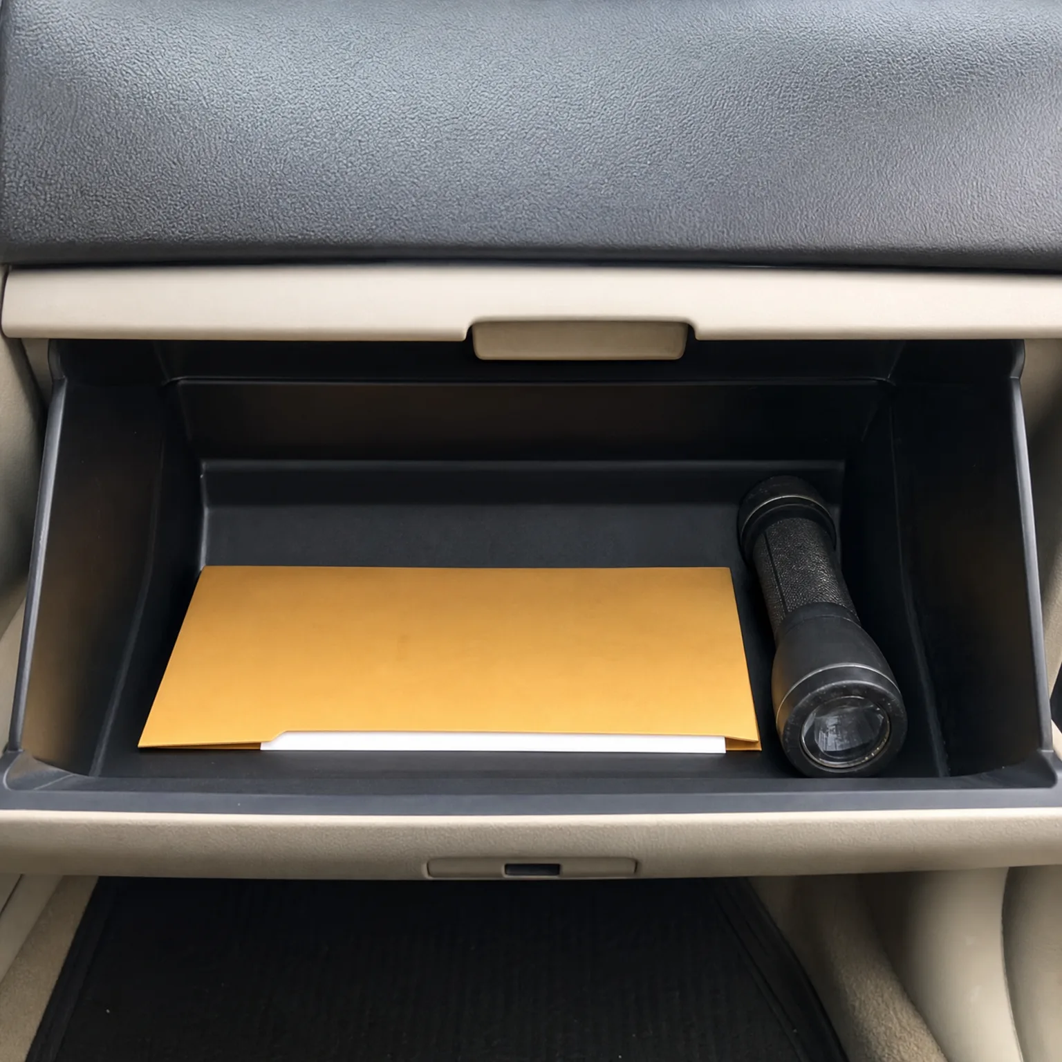 An open car glove compartment containing a yellow envelope over white papers and a black flashlight.