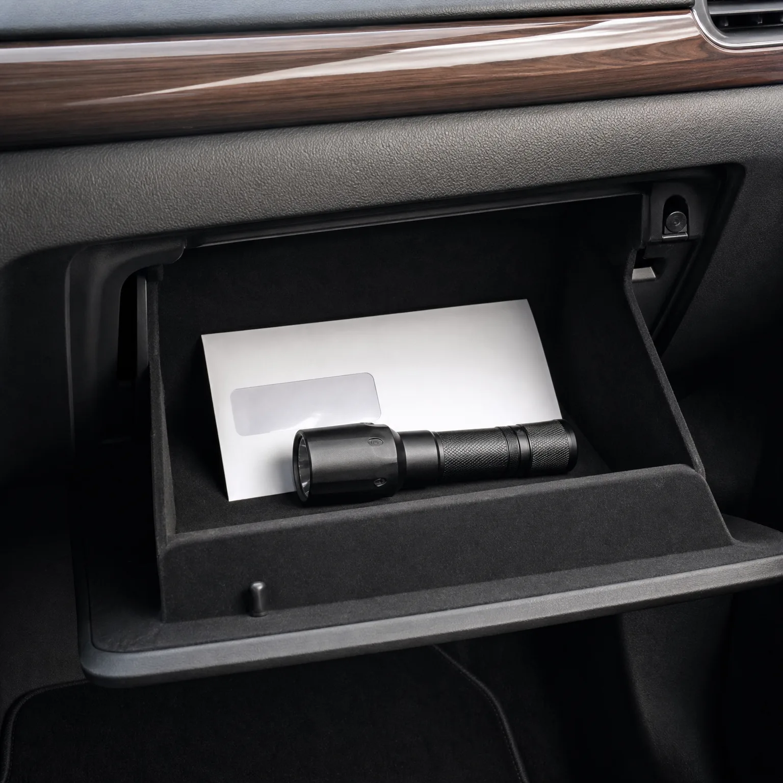 An open car glove compartment containing a black flashlight resting on a white booklet, with wood-grain dashboard trim above.