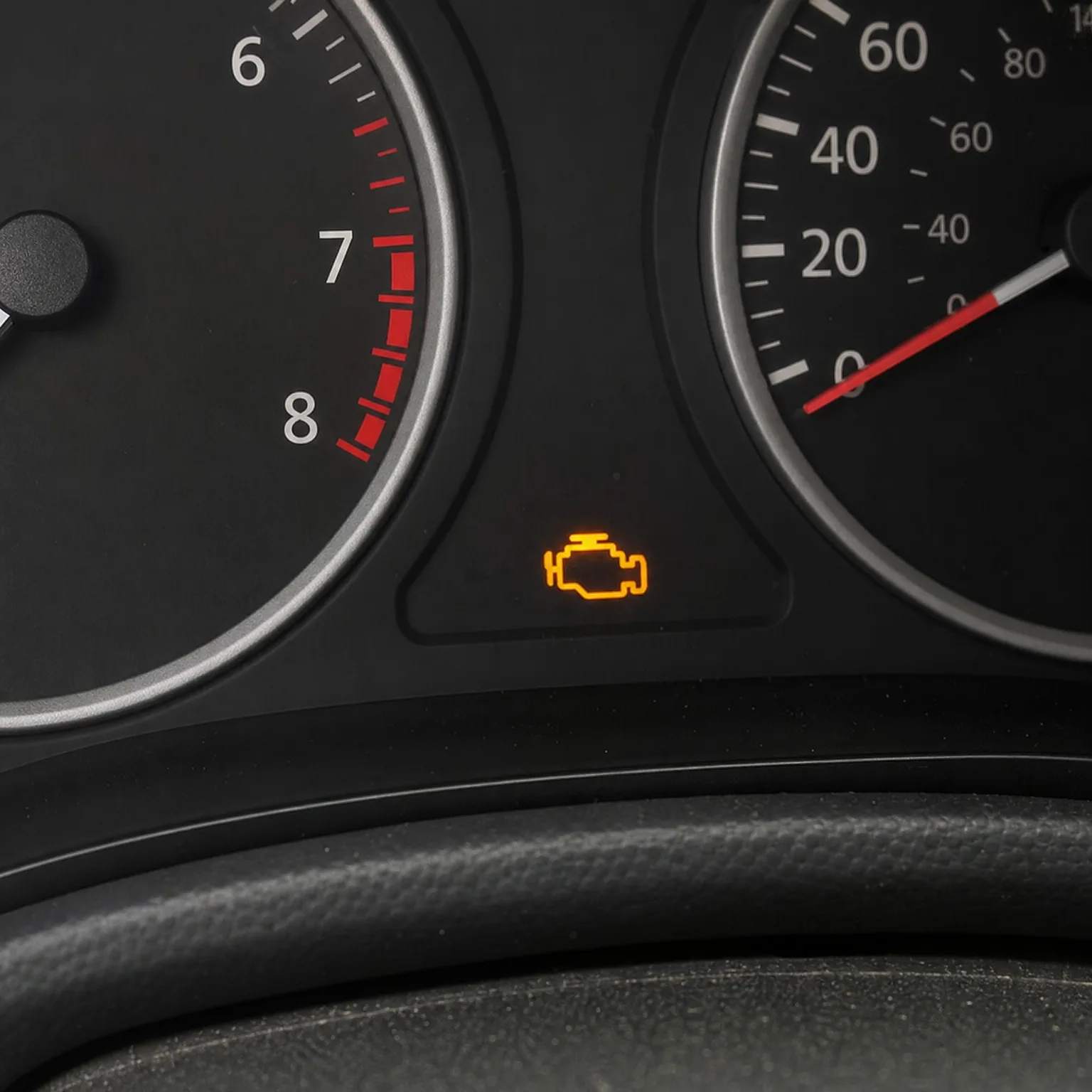 Close-up of a car instrument cluster showing a tachometer with red zone near 7-8 and a speedometer, with an illuminated orange engine-shaped warning light between them.