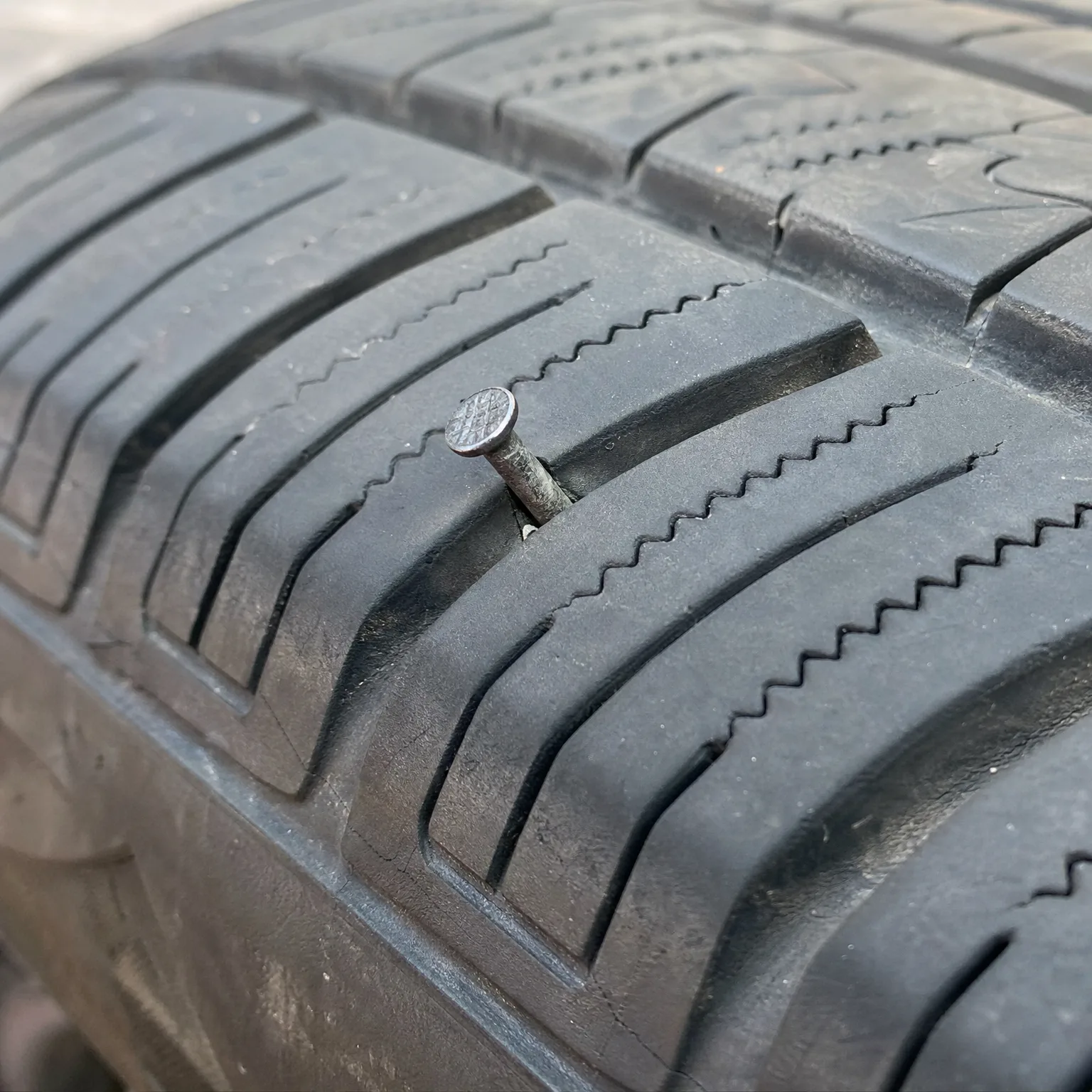 Close-up of a black tire tread with a nail embedded in a groove between tread blocks.