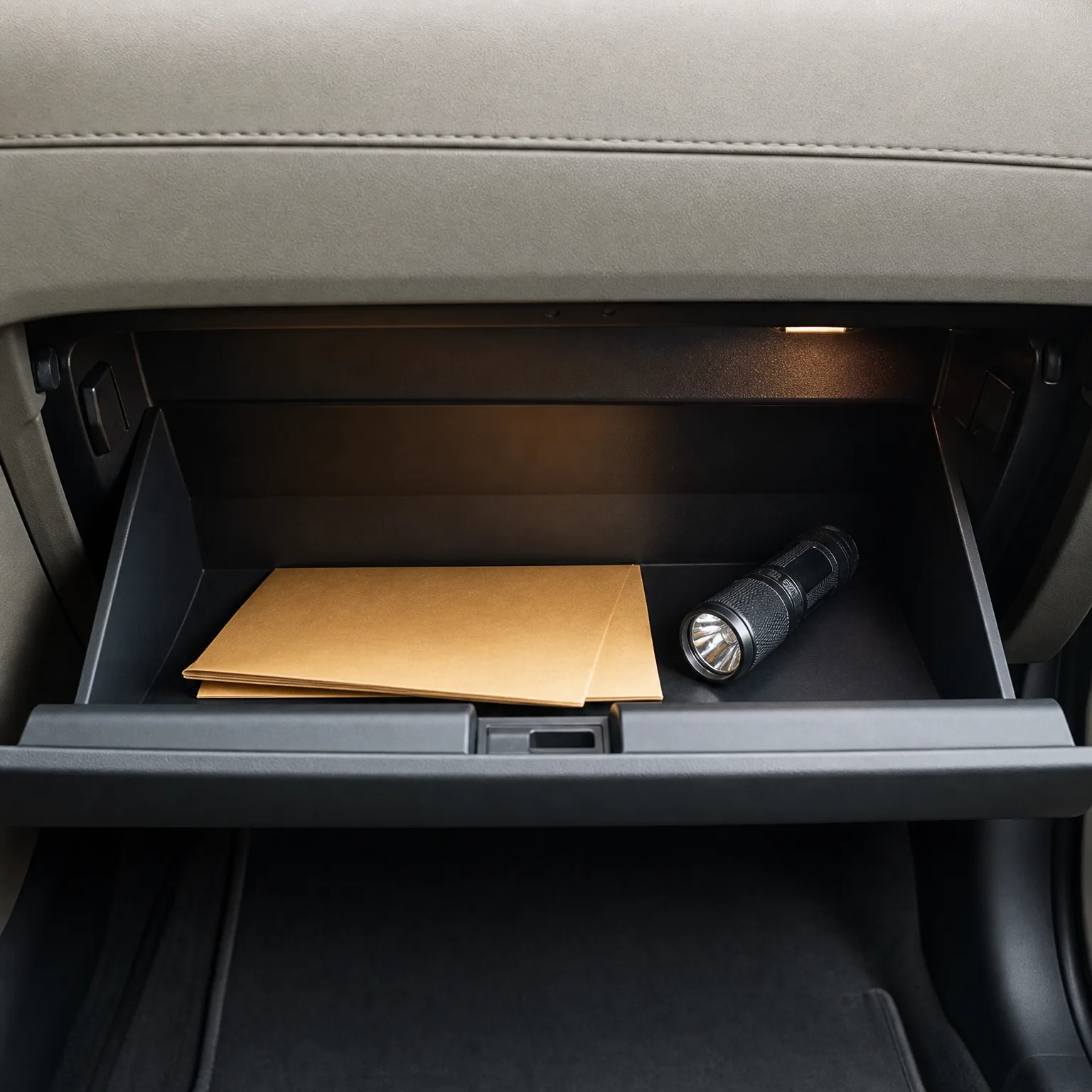 An open car glove compartment containing a stack of brown paper envelopes and a black flashlight, with an interior light illuminating the compartment.