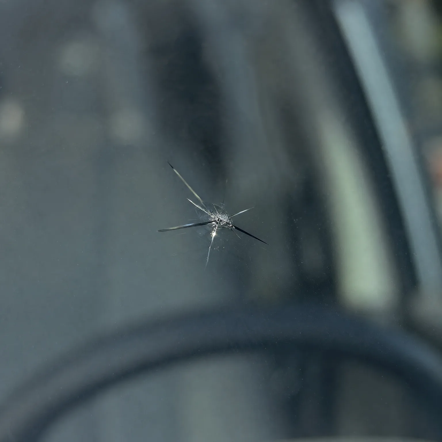 Close-up of a star-shaped chip with radiating cracks in a vehicle windshield, with a blurred dark interior and side window behind.