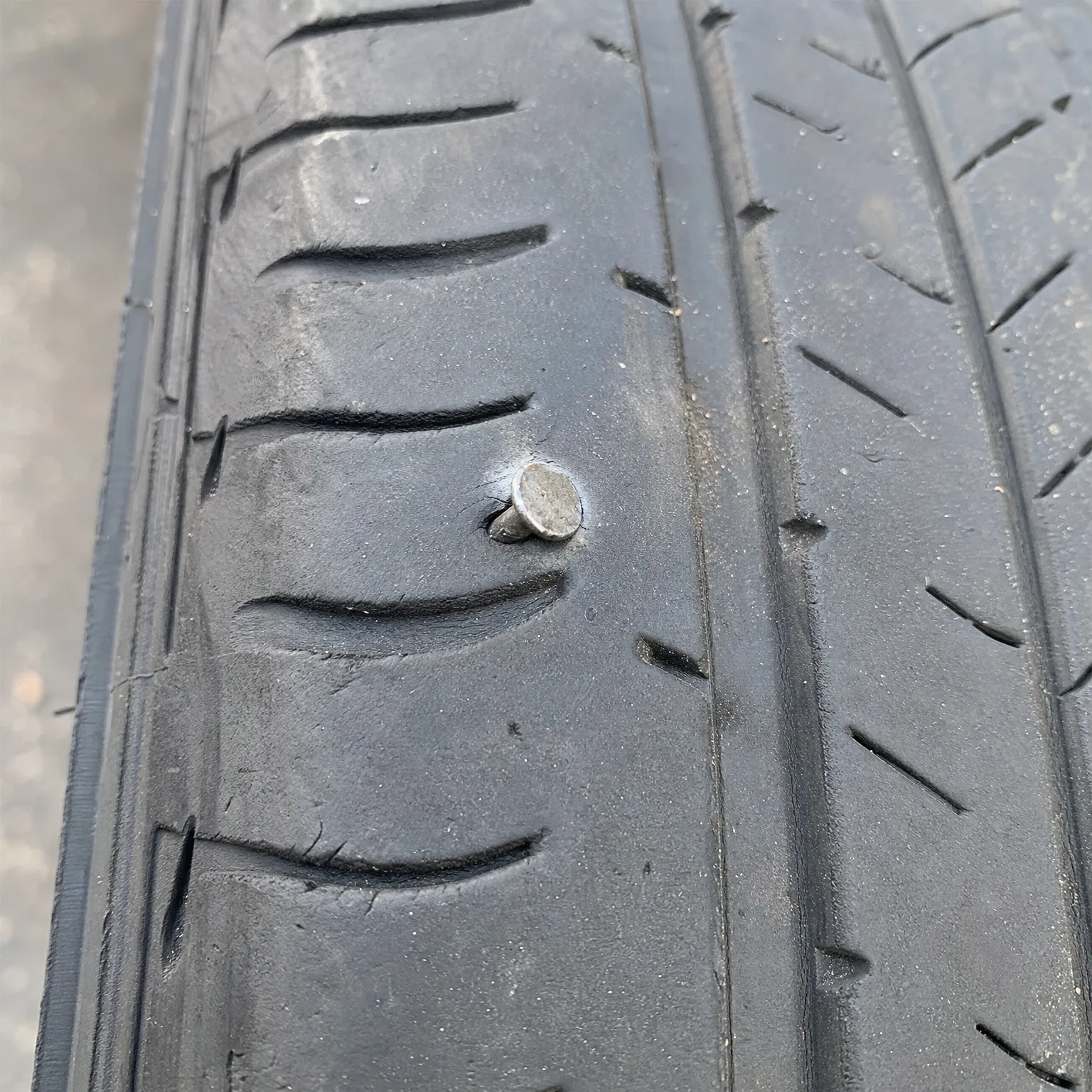 Close-up of a car tire tread with a metal nail head embedded in the rubber between grooves.