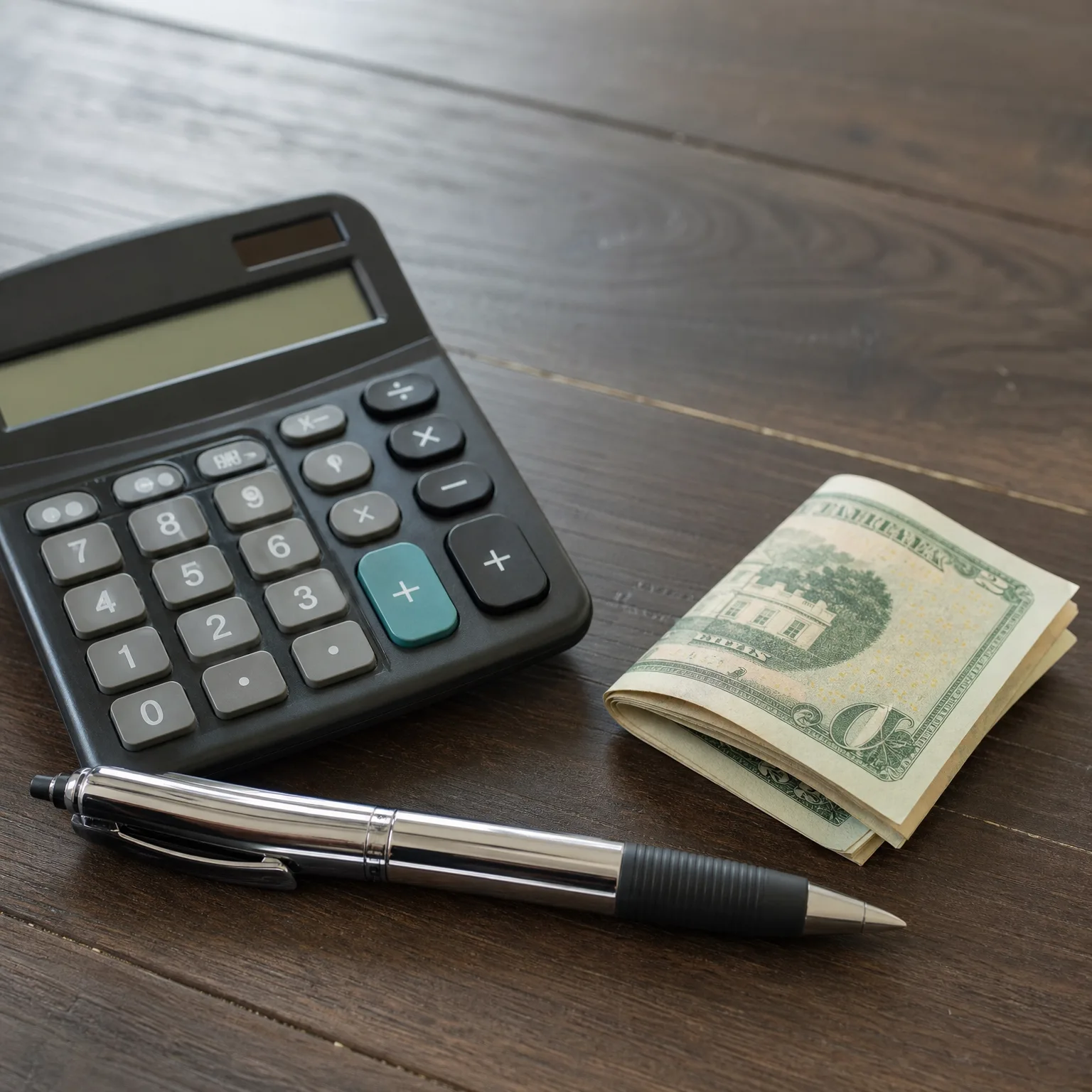 A black desktop calculator, a silver ballpoint pen, and a folded stack of banknotes on a dark wooden surface.