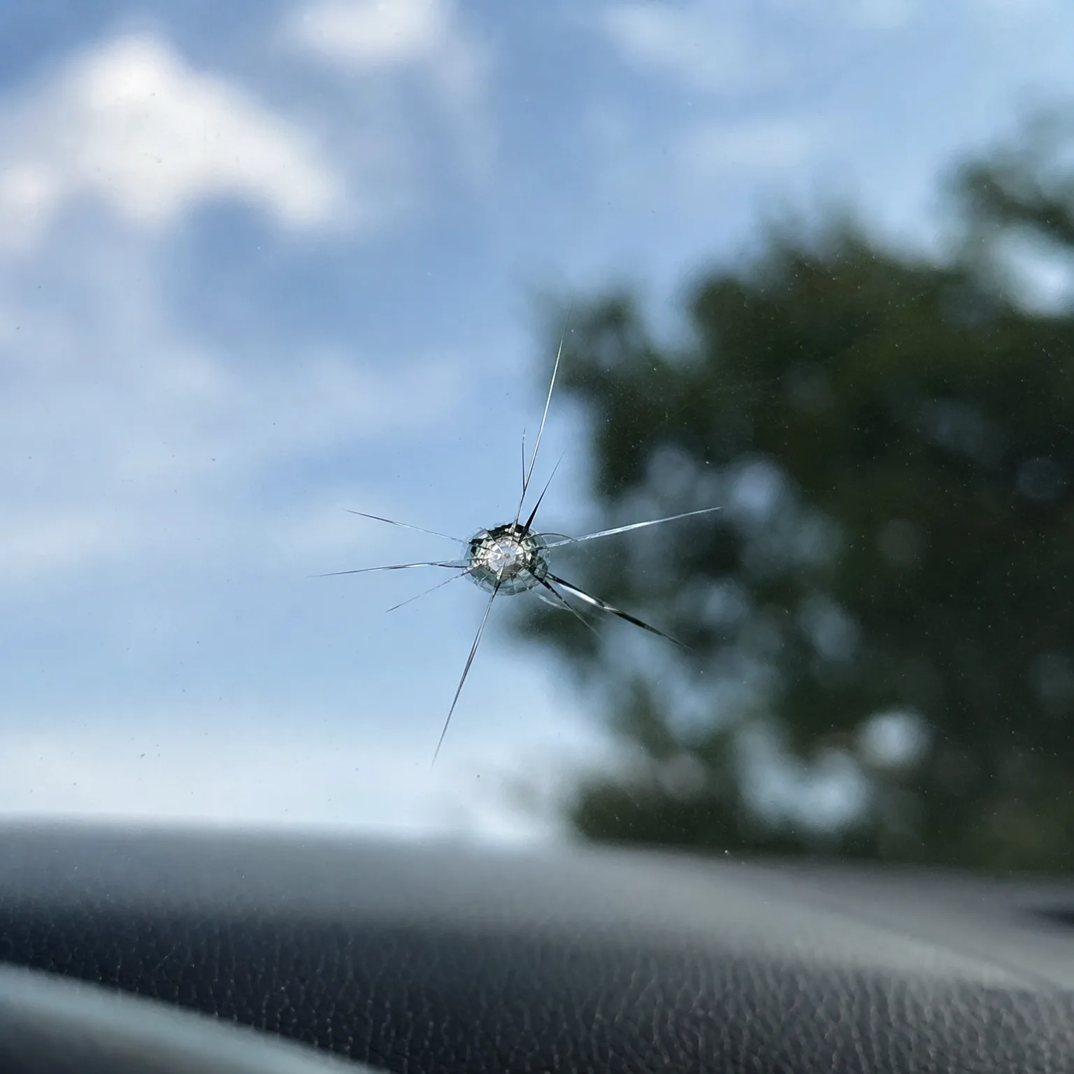 A star-shaped chip with radiating cracks in a car windshield, seen against a blue sky with clouds and a blurred tree, with the dark dashboard below.