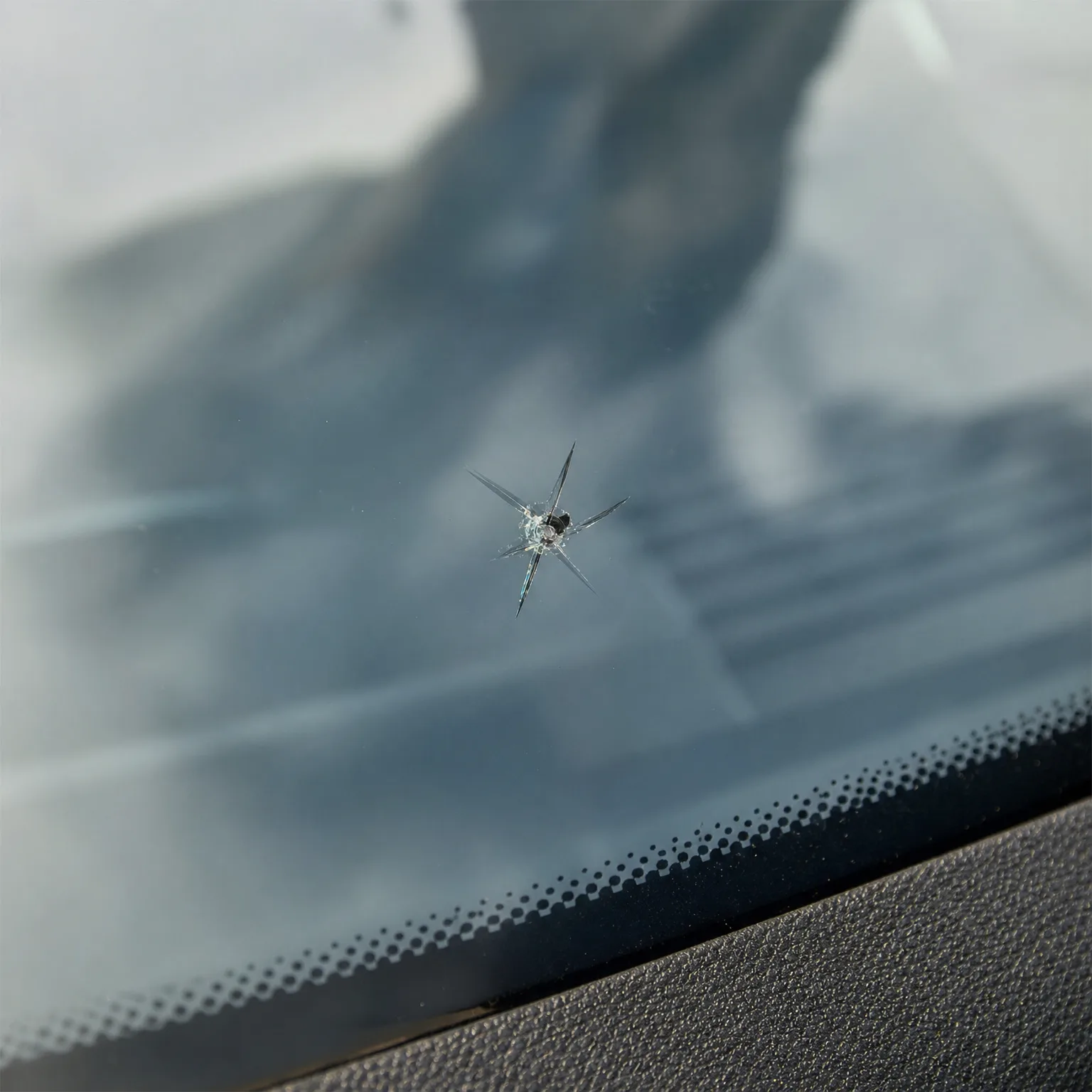 Close-up of a star-shaped chip with radiating cracks in a car windshield, with the dotted black frit border and dashboard edge visible at lower right.
