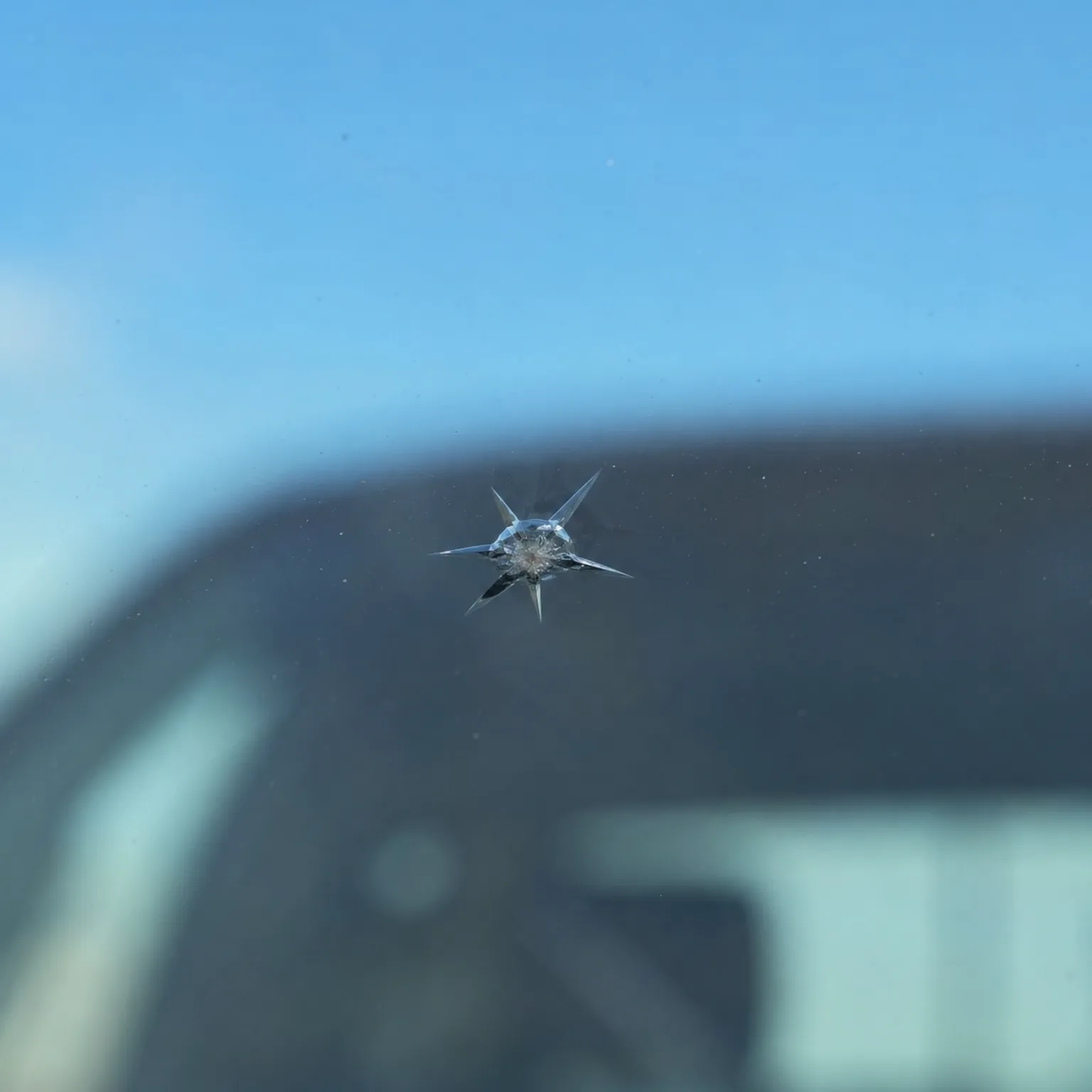 Close-up of a star-shaped chip in a vehicle windshield, with blurred blue sky and dark car structure in the background.