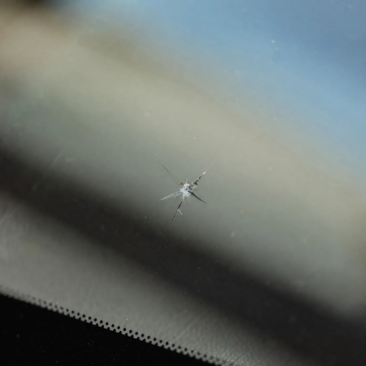 Close-up of a star-shaped chip crack in a vehicle windshield, with a blurred dashboard and defroster vents below and sky above.