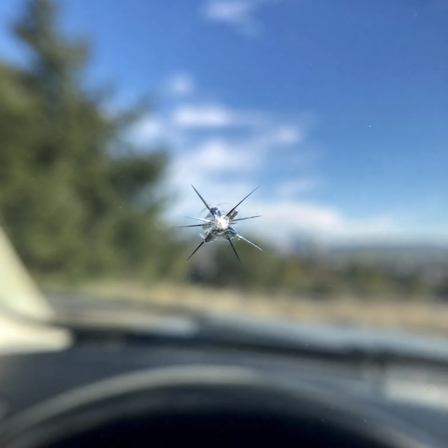 A star-shaped chip with radiating cracks in a vehicle windshield, seen from inside the car against a blue sky with scattered clouds, blurred evergreen trees and a dark dashboard.