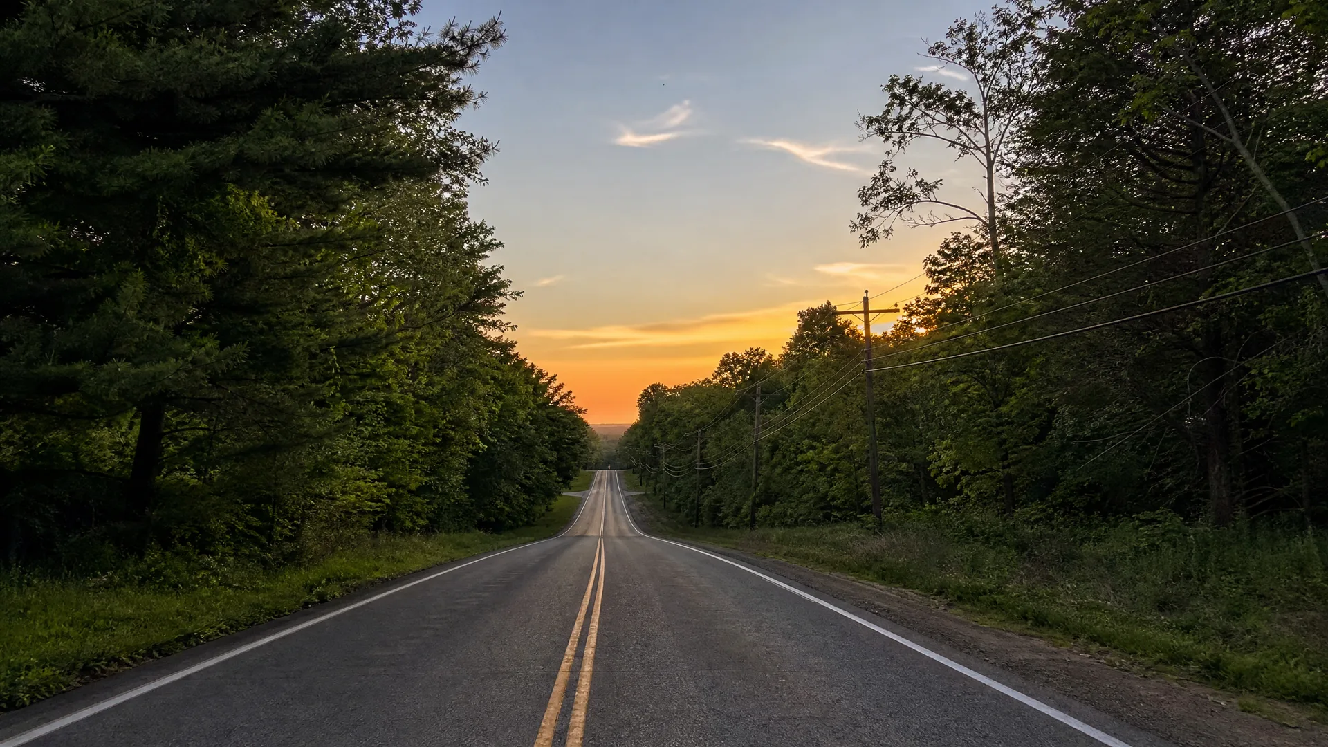 A two-lane asphalt road with double yellow center lines runs straight toward a dipping horizon, flanked by dense green trees and utility poles with wires, under an orange and blue sky at sunset.