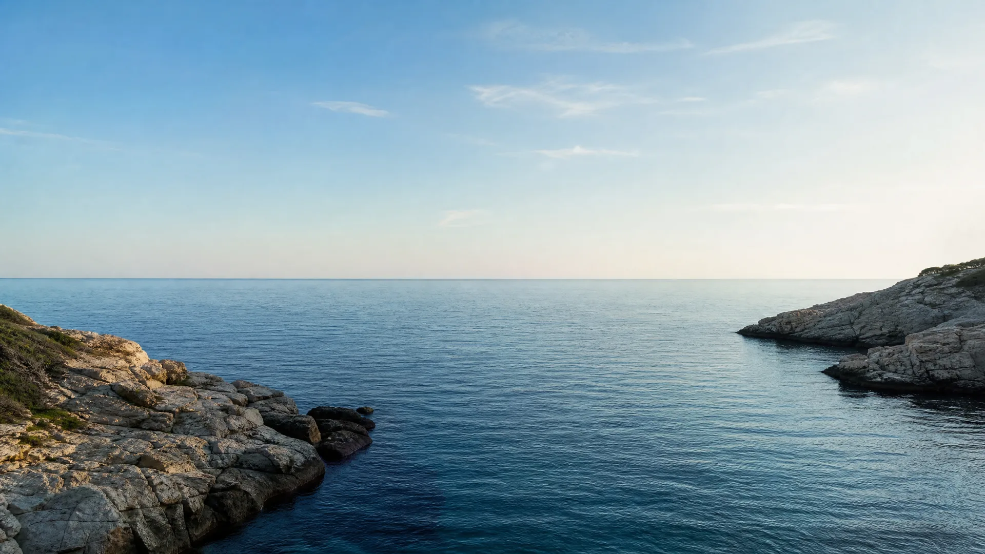 Calm blue sea under a clear pale sky, framed by low rocky limestone shores with sparse scrub in the lower left and right foreground.
