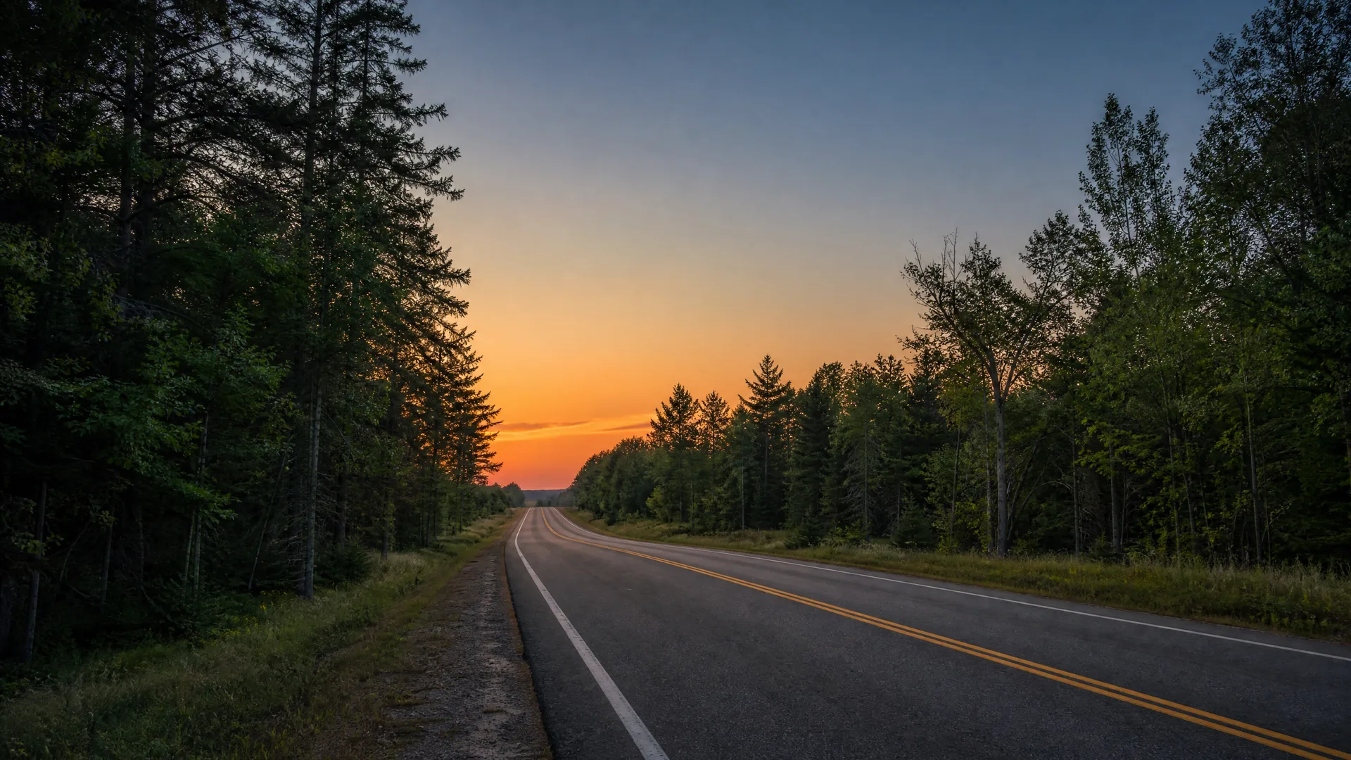 A two-lane asphalt road with double yellow center lines curves through dense conifer and deciduous forest toward an orange sunset sky.