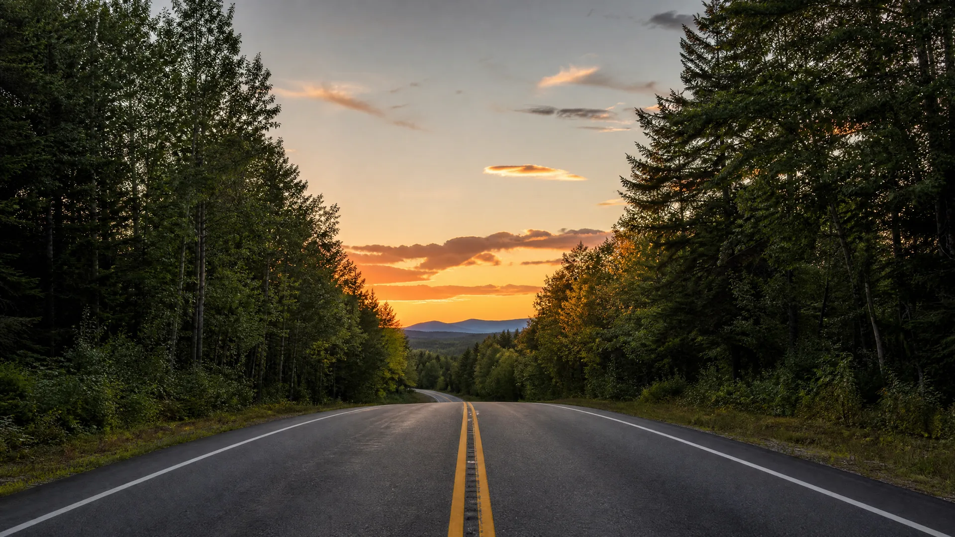 A two-lane paved road with double yellow center lines runs toward distant hills, lined by dense conifer forest under an orange sunset sky.