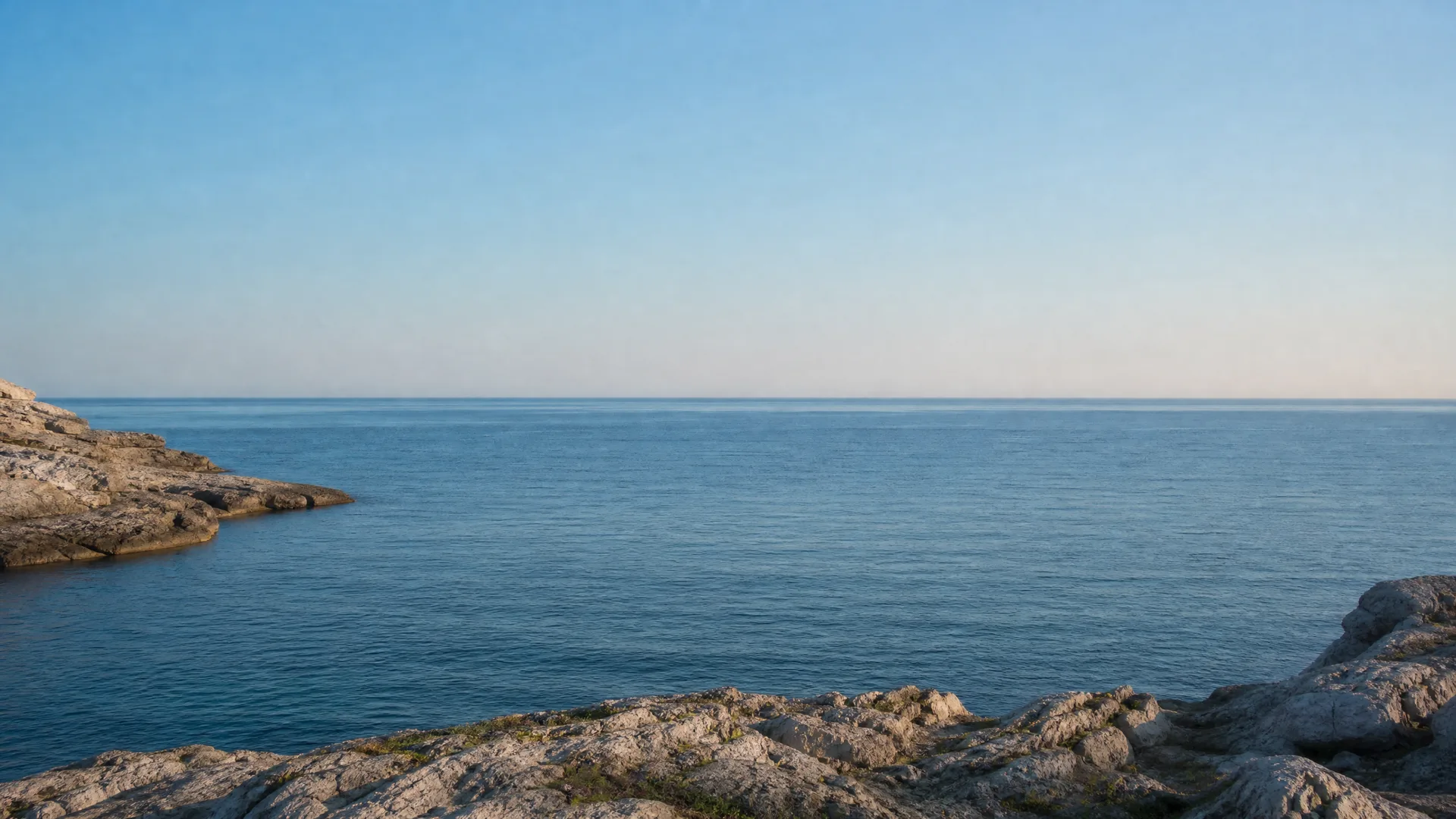 A calm sea under a clear pale blue sky, framed by rocky shoreline in the foreground and at the left edge.
