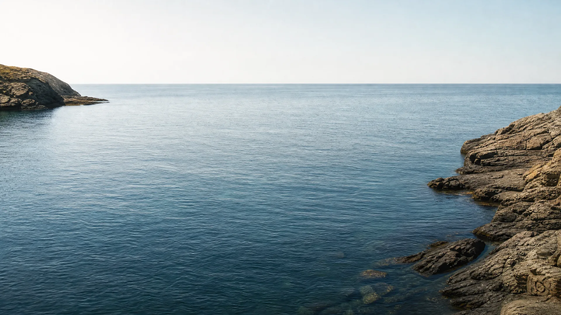 Calm blue sea under a clear pale sky, framed by rocky shoreline outcrops at the right and a low headland at the far left.