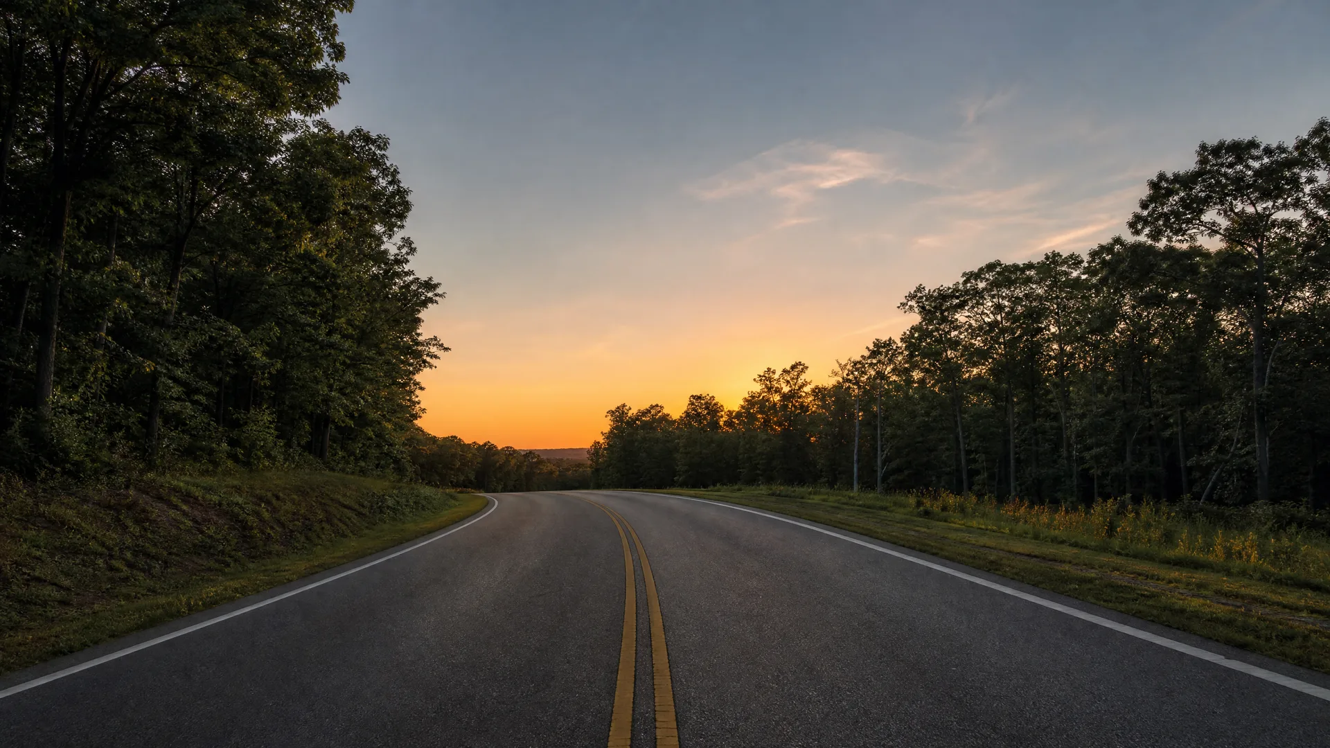 A two-lane asphalt road with a double yellow center line curving left between dense deciduous trees, under an orange and blue sky at dusk.