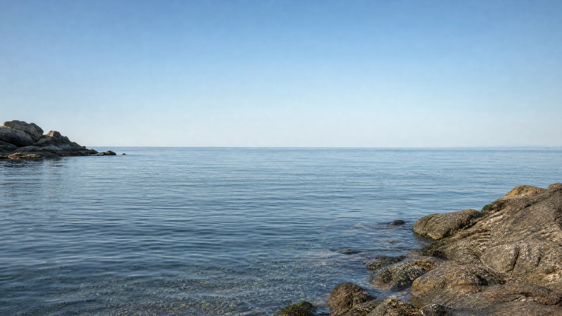 Calm blue sea under a clear sky, with rocky outcrops in the lower right foreground and a low rocky point on the left horizon.