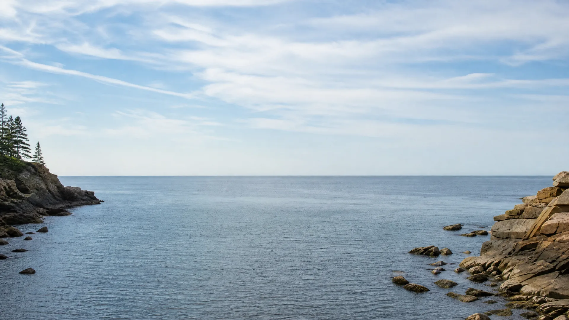 A calm ocean cove framed by rocky headlands, with conifers on the low cliff at left and tan boulders at right under a mostly clear sky with thin clouds.