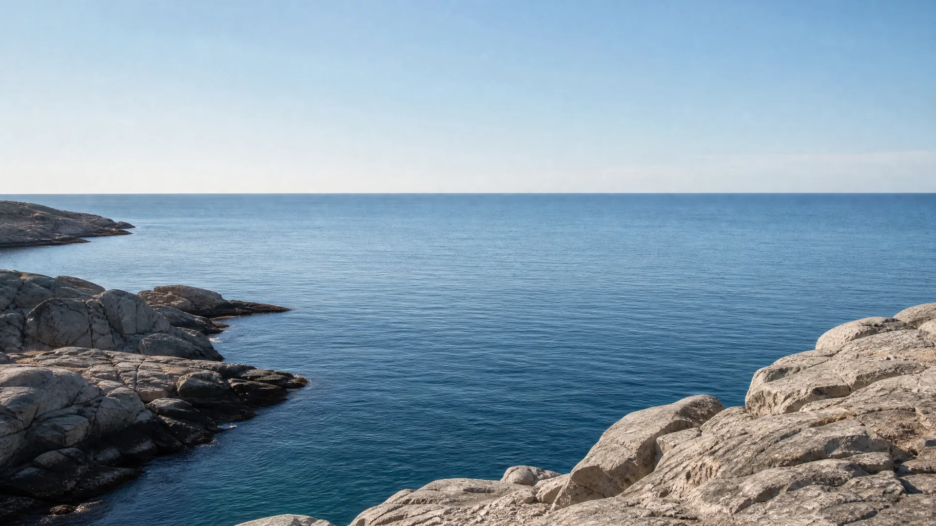 Smooth grey rocky shoreline in the foreground and left side framing calm blue sea under a clear pale sky with a flat horizon.