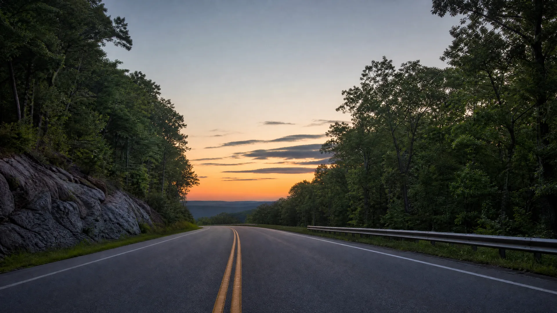 A two-lane road with double yellow center lines curves toward distant hills at dusk, bordered by a rock cut and trees on the left and a guardrail with dense trees on the right under an orange and blue sky.