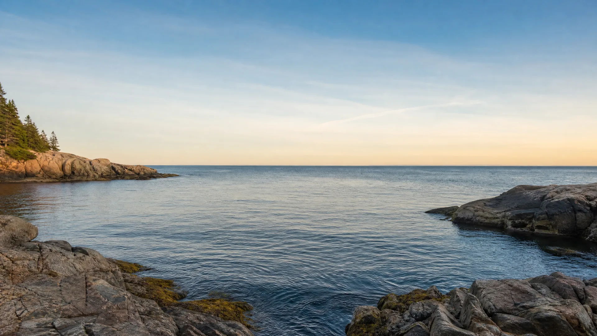 A calm sea in a rocky cove at sunrise or sunset, with granite ledges in the foreground and right, and a low wooded headland with conifers on the left.