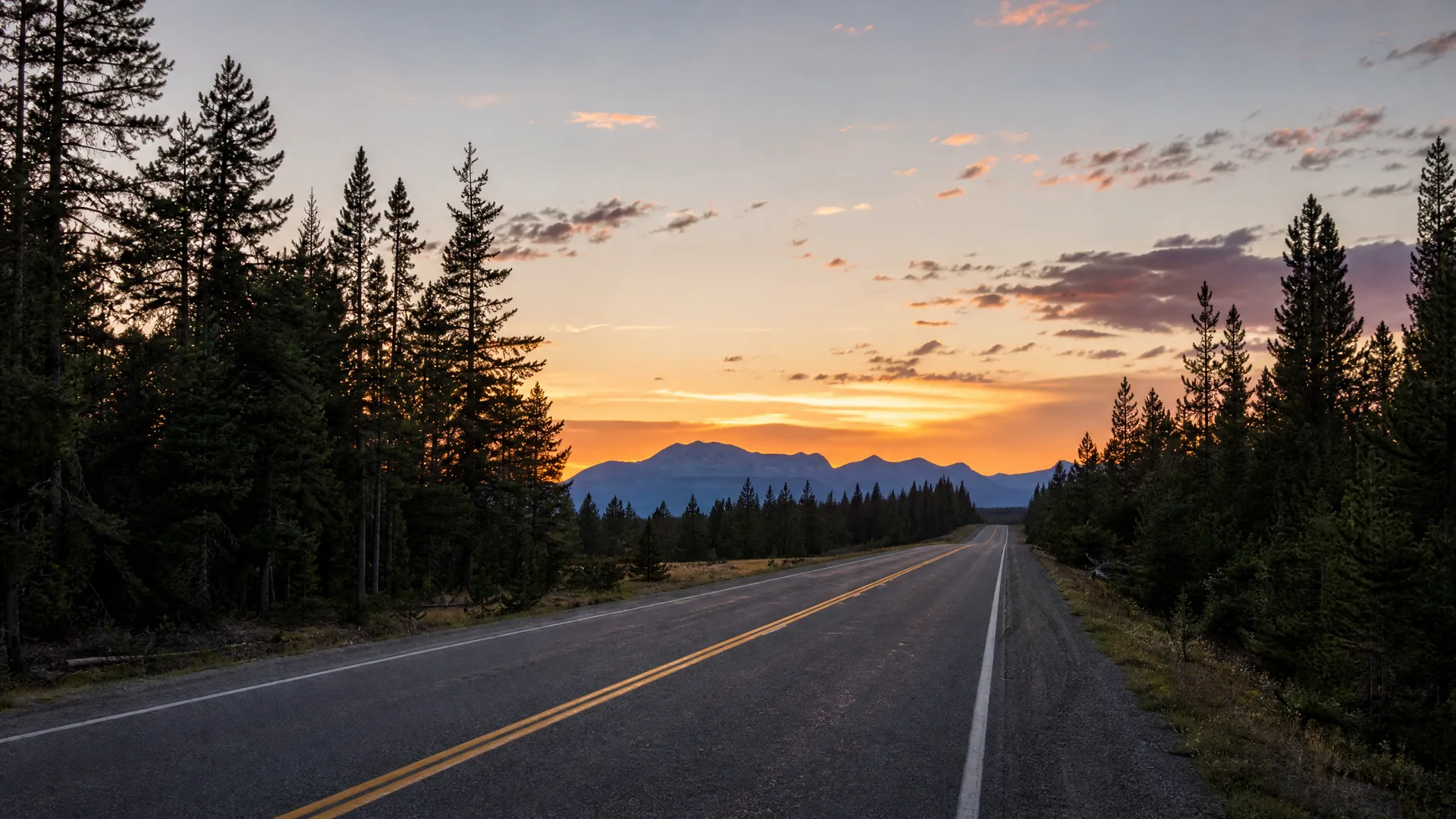 A two-lane road with double yellow center lines runs toward distant mountains, flanked by conifer forest under an orange and purple sunset sky.