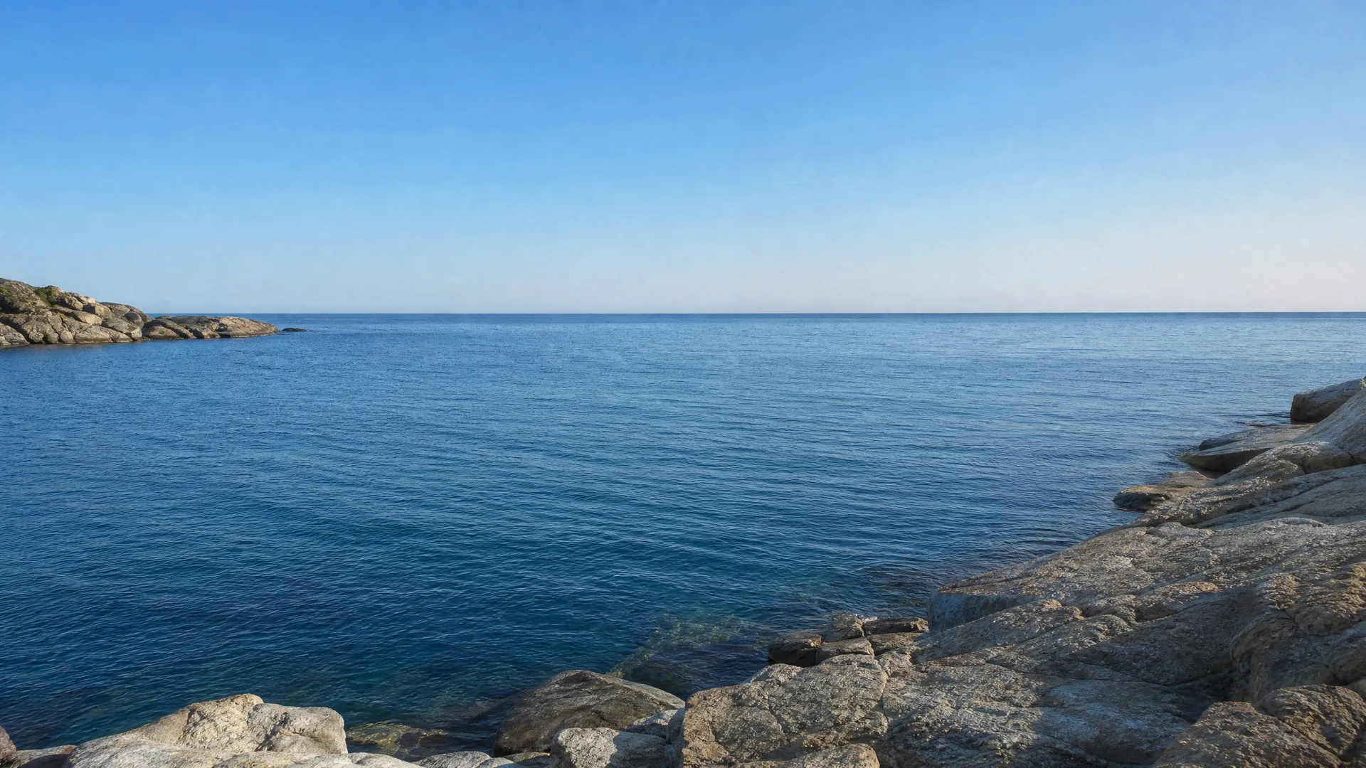 A calm blue sea under a clear sky, bordered by pale rocky outcrops in the foreground right and a low rocky point on the left.